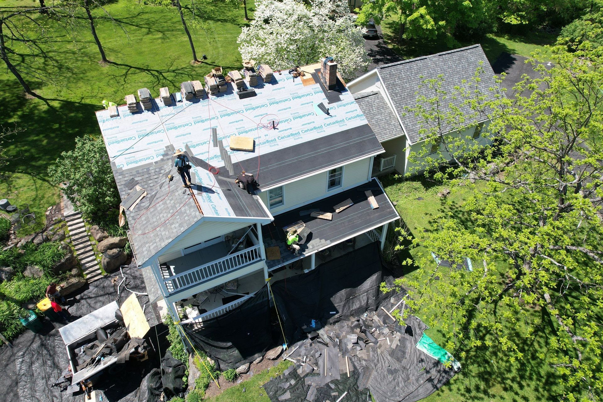 Overhead view of a house undergoing roof replacement. Workers on roof, supplies laid out. Green trees surround.