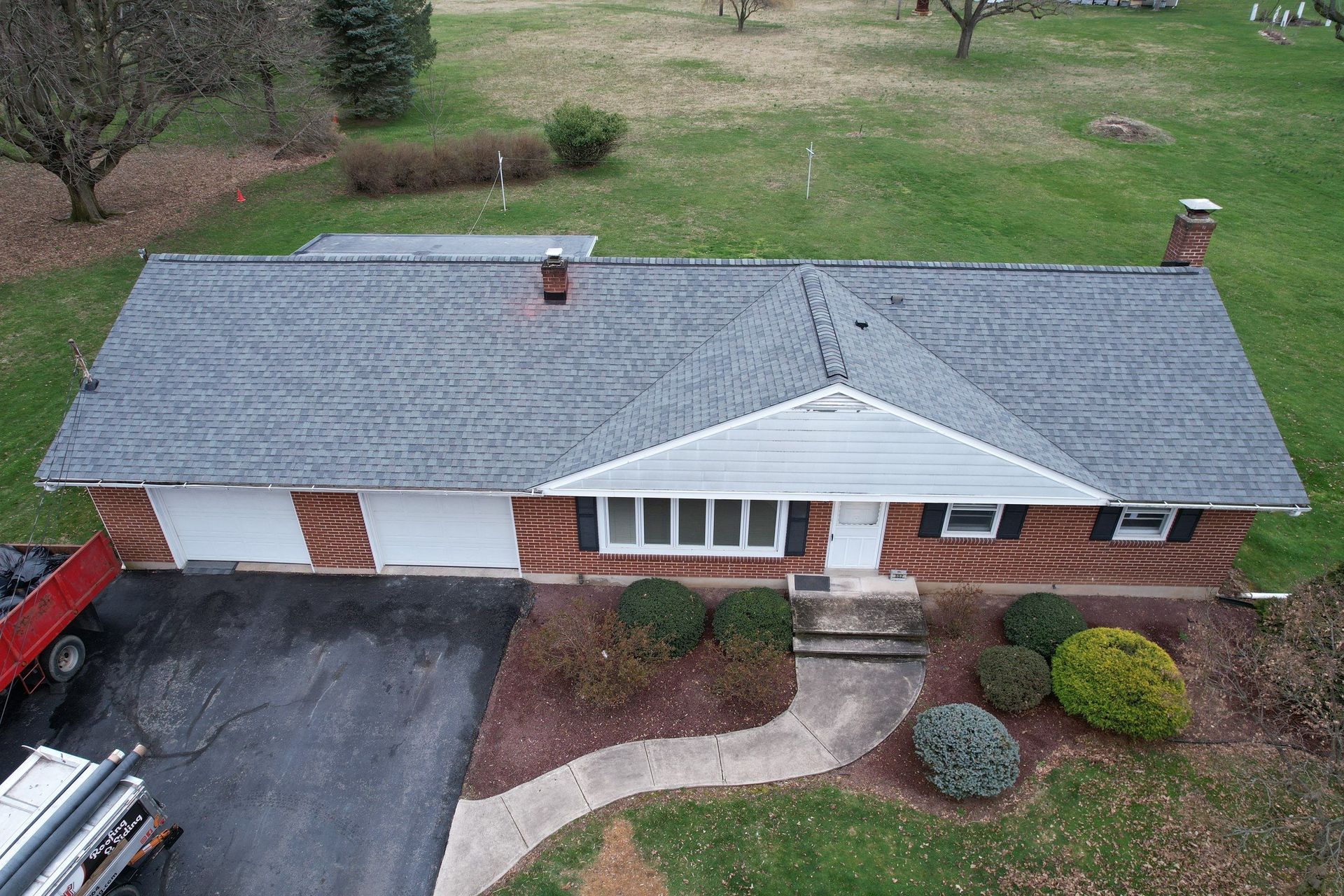Aerial view of a single-story brick house with a gray roof and two-car garage, set on a green lawn with landscaping.