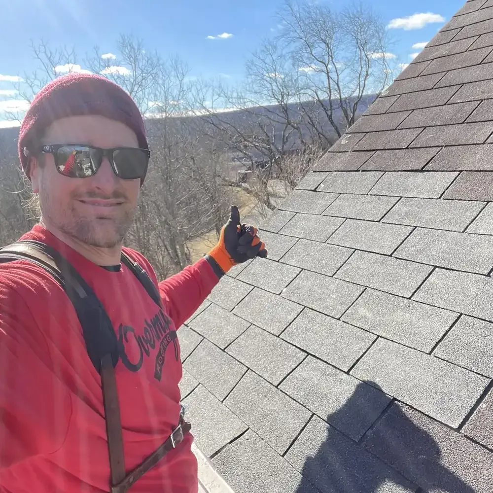 A man wearing sunglasses and a red shirt is standing on top of a roof giving a thumbs up.