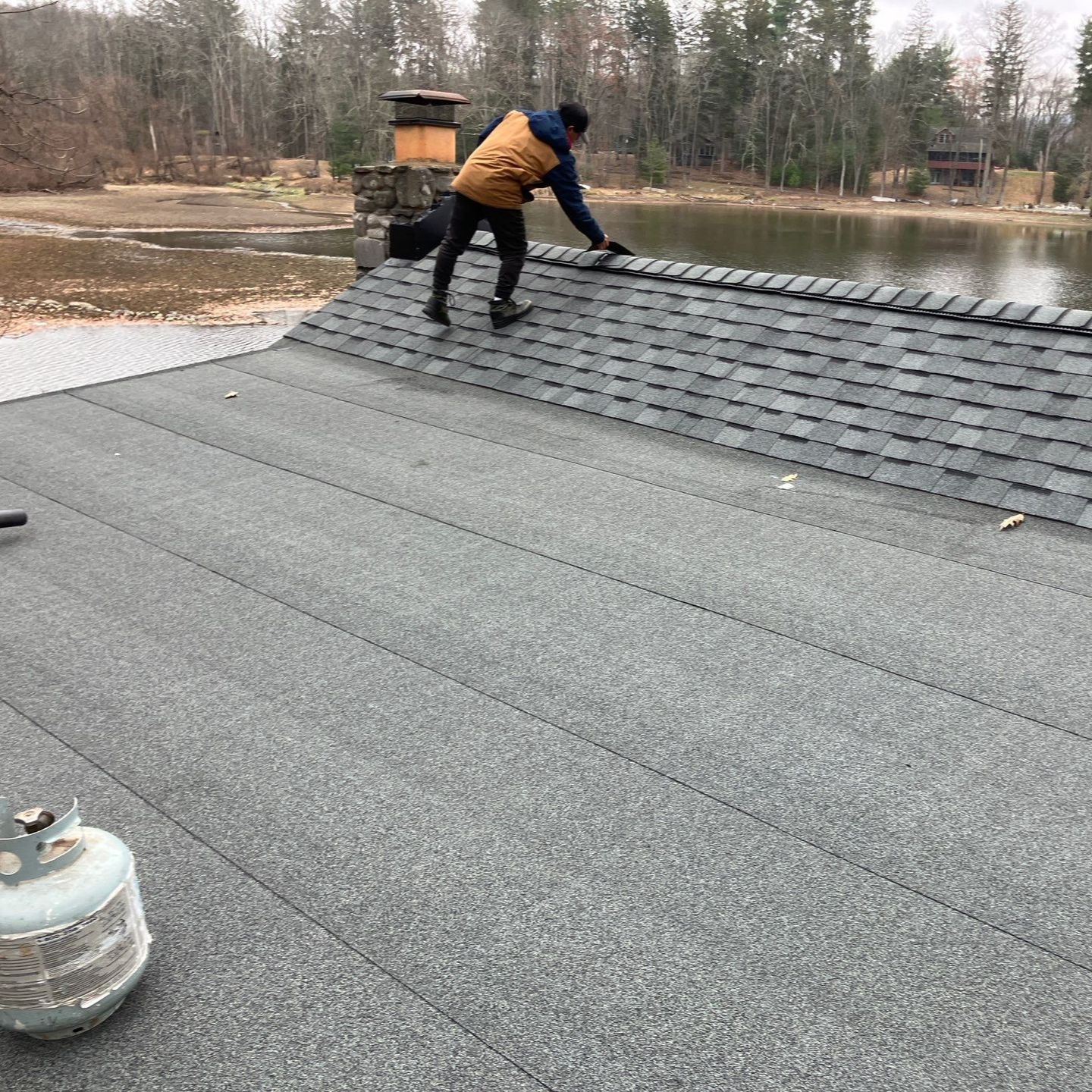 Worker crouching on a gray roof near a lake, installing shingles outdoors. Worker crouching on a gray roof near a lake, installing shingles outdoors.