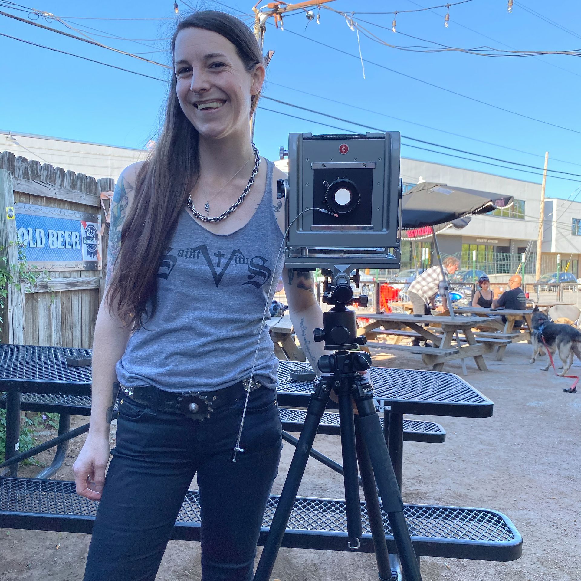 Woman standing beside a large camera on a tripod at an outdoor venue with picnic tables and people in the background