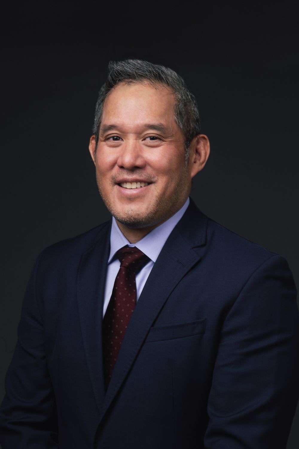 A smiling person wearing a dark blue suit, light blue dress shirt, and a maroon tie against a dark grey background.