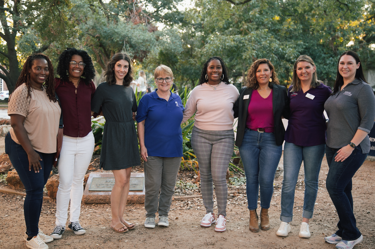 A group of eight people stand in a line outdoors in a park setting, smiling for a group portrait.