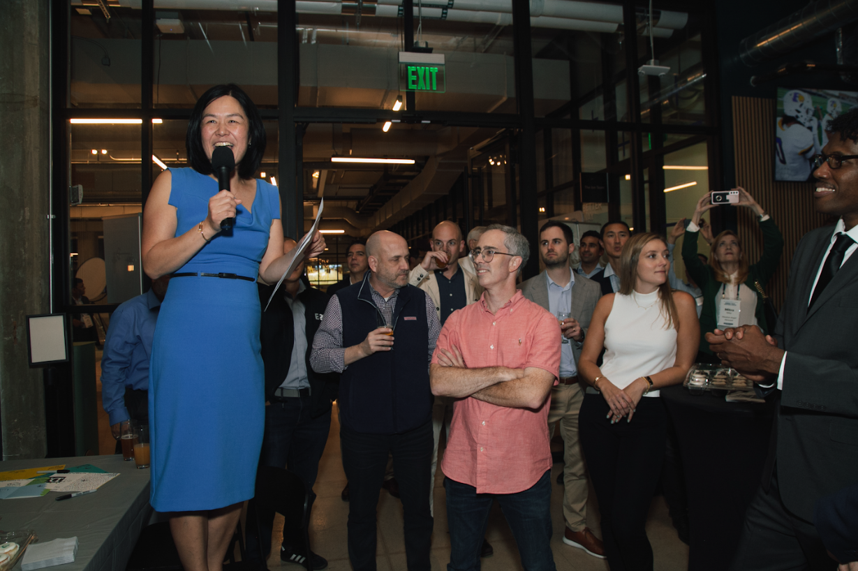 A person in a blue dress speaks into a microphone at a gathering with several attendees listening in an office space.