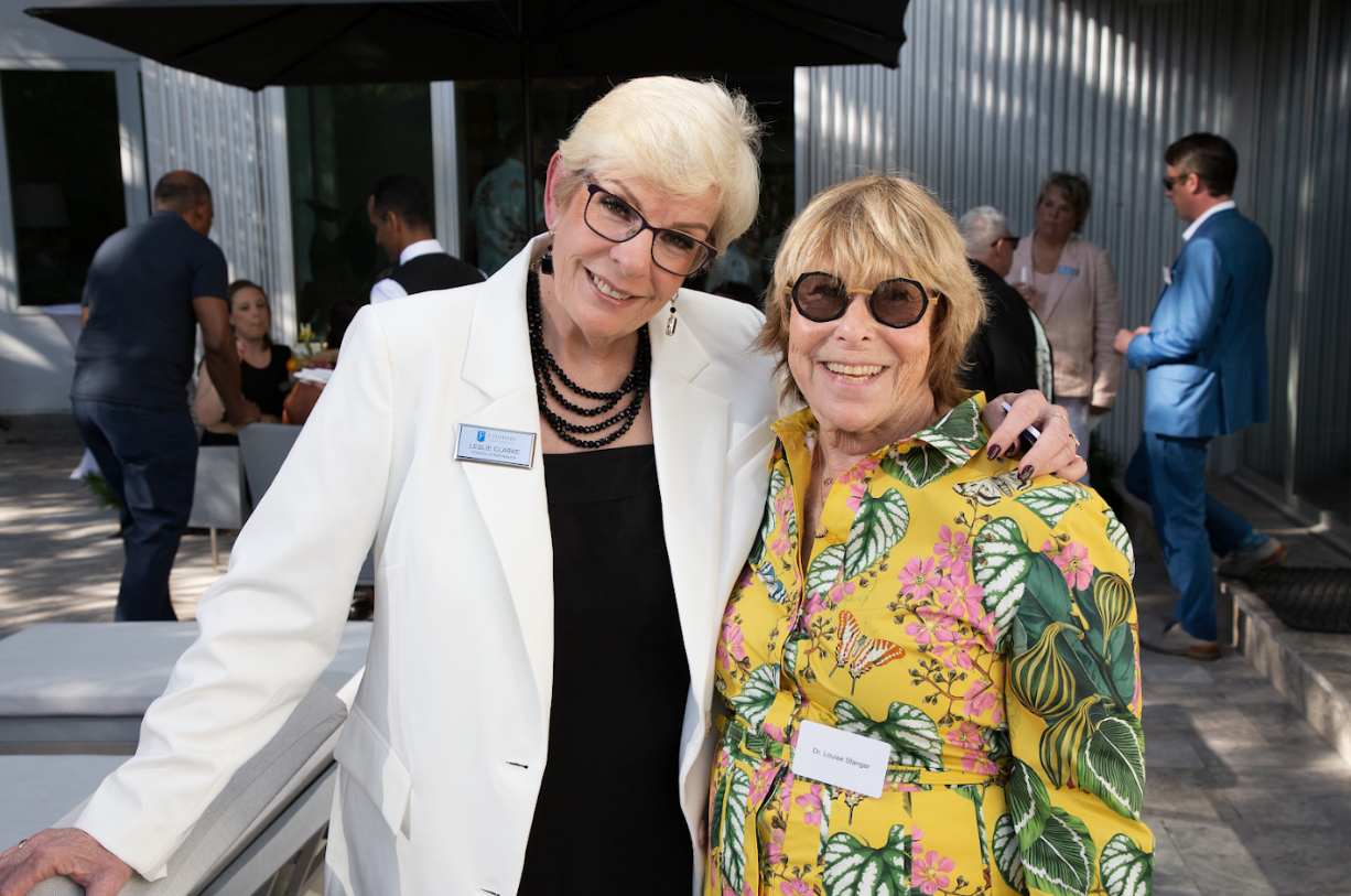 Two people stand arm-in-arm at an outdoor event, one in a white blazer and black top, the other in a yellow floral dress.