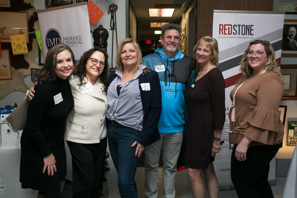Six people stand smiling together for a group photo at an indoor event with branded banners in the background.