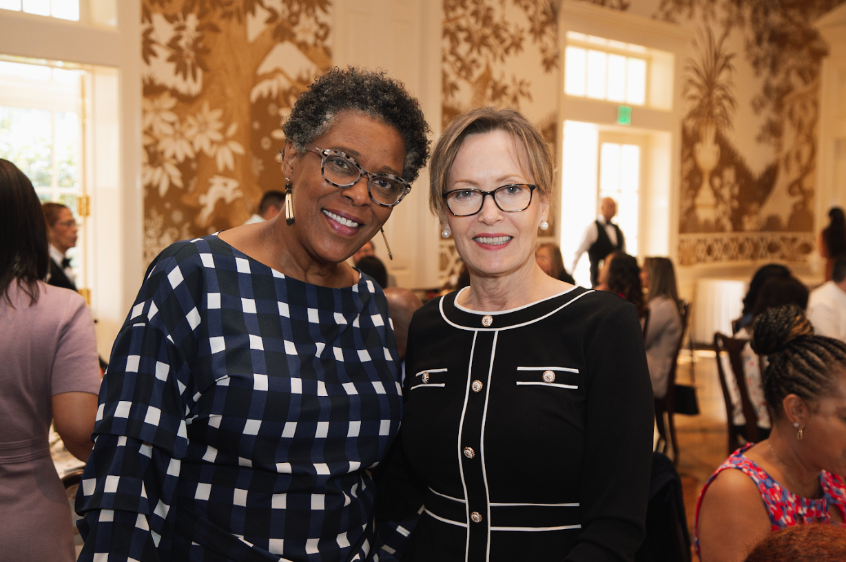 Two people in glasses smile together at an indoor event with patterned wallpaper in the background.