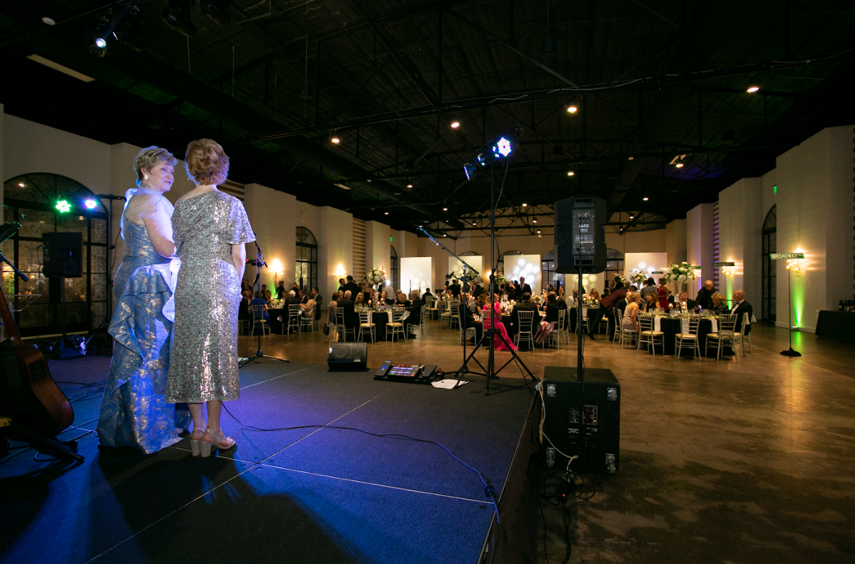 Two performers in formal attire stand on a stage facing a seated audience in a large, dimly lit banquet hall.