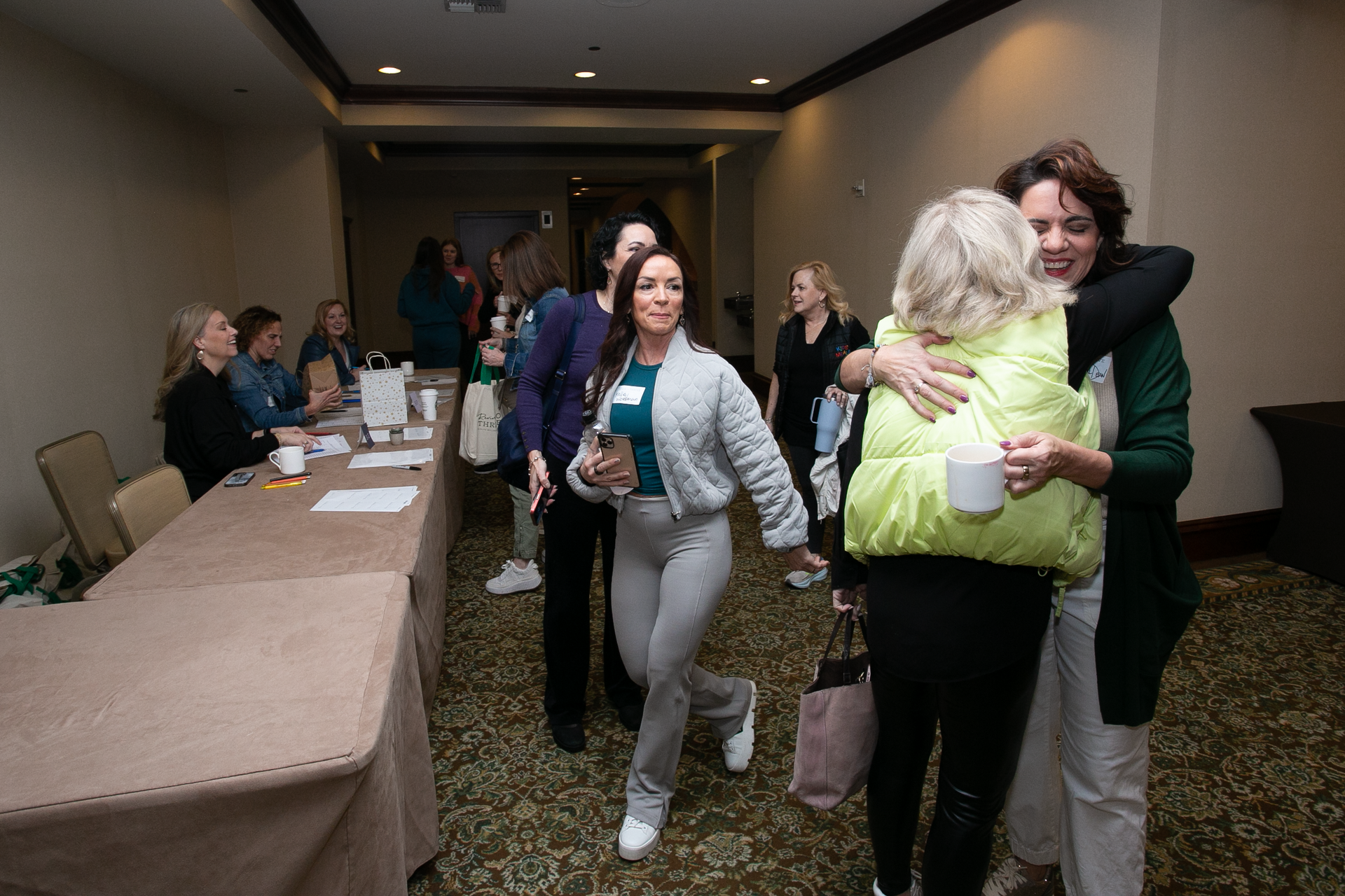 Two people embrace in a hotel conference area while others gather at tables in the background.