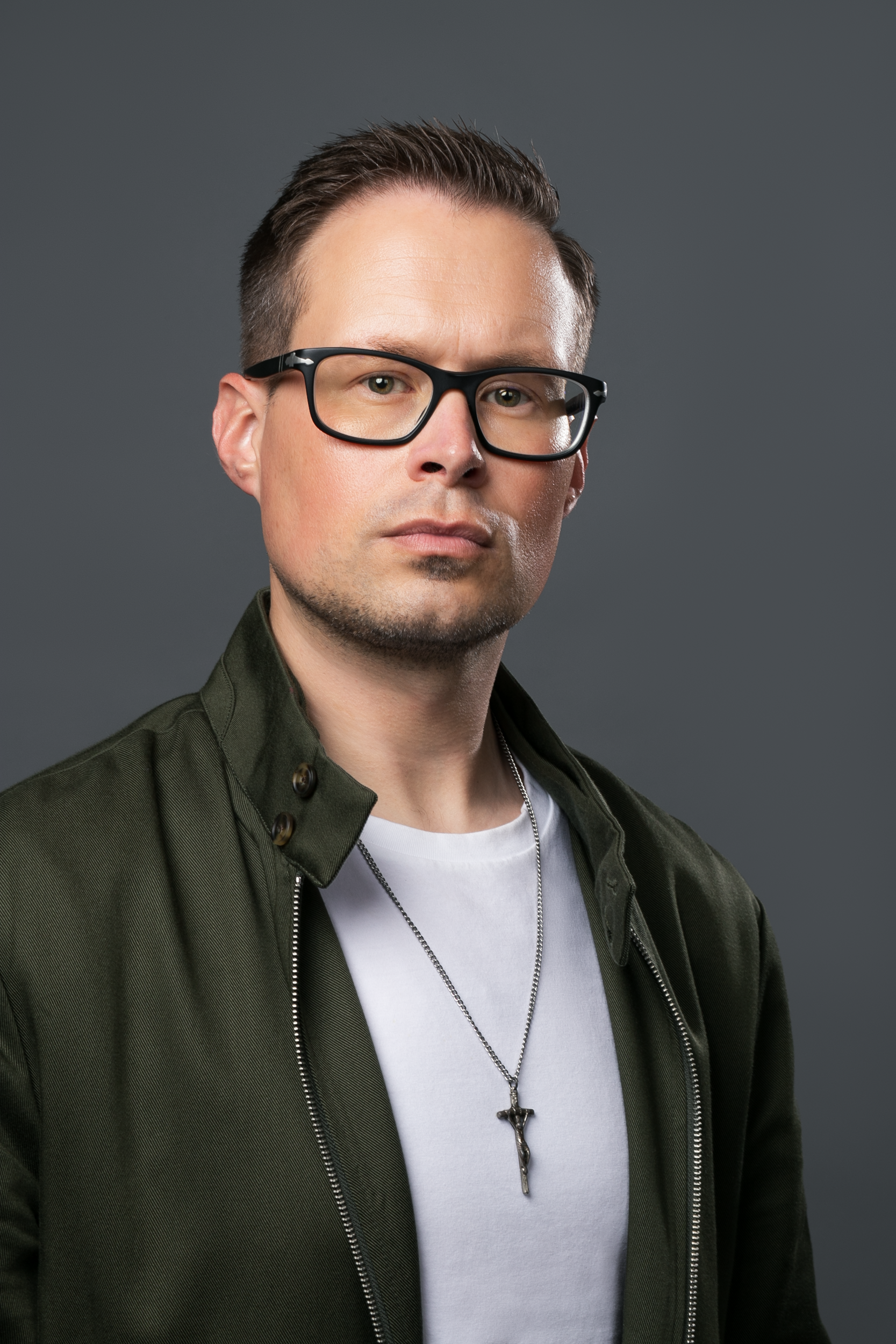A person wearing glasses, a white shirt, a dark jacket, and a cross necklace, posed against a gray background.