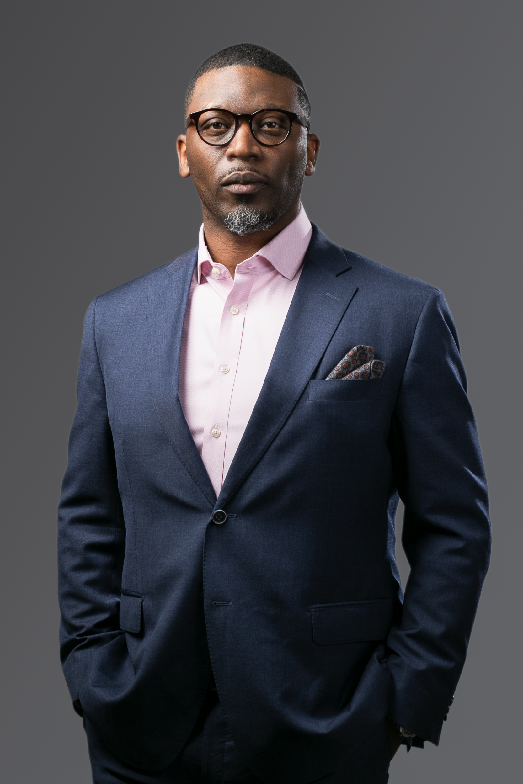 A professional portrait of a person wearing glasses, a pink dress shirt, and a dark blue blazer against a grey background.
