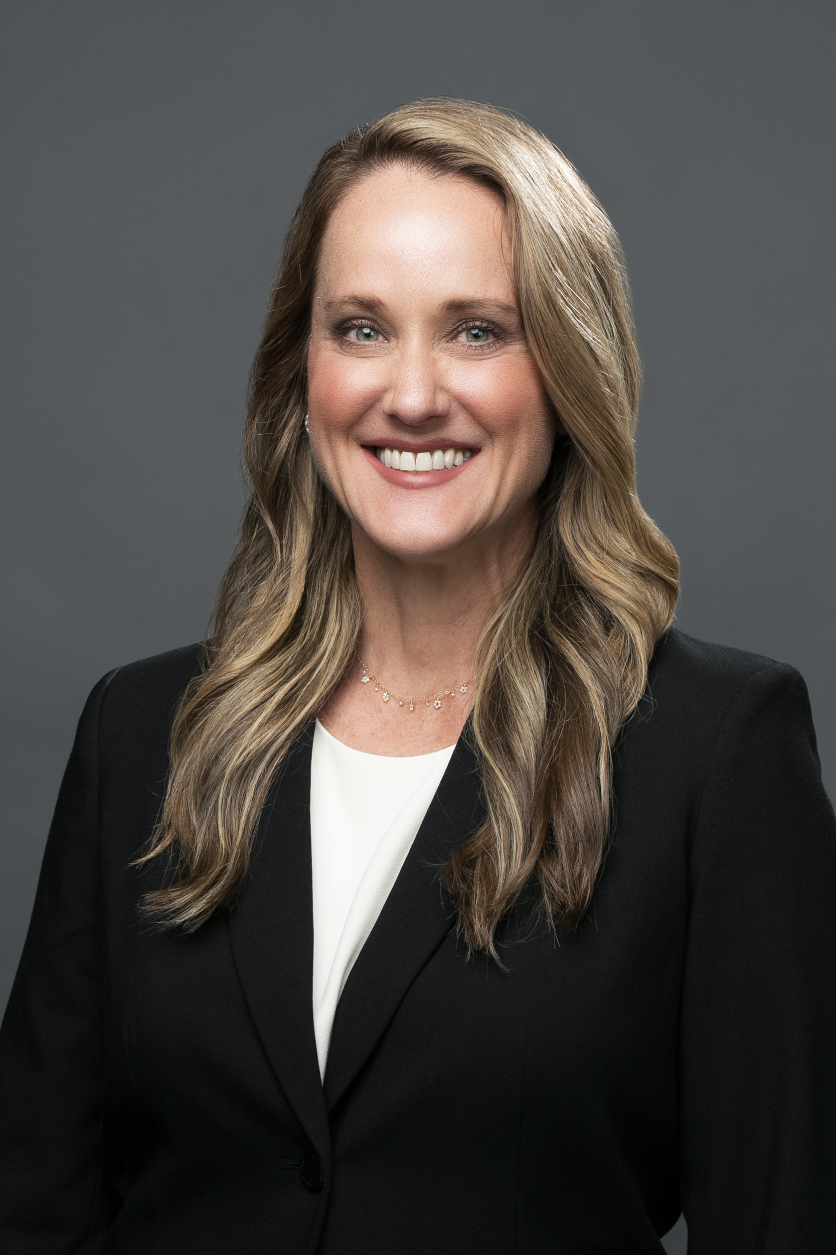A smiling professional person with long wavy hair, wearing a white shirt and black blazer against a grey background.