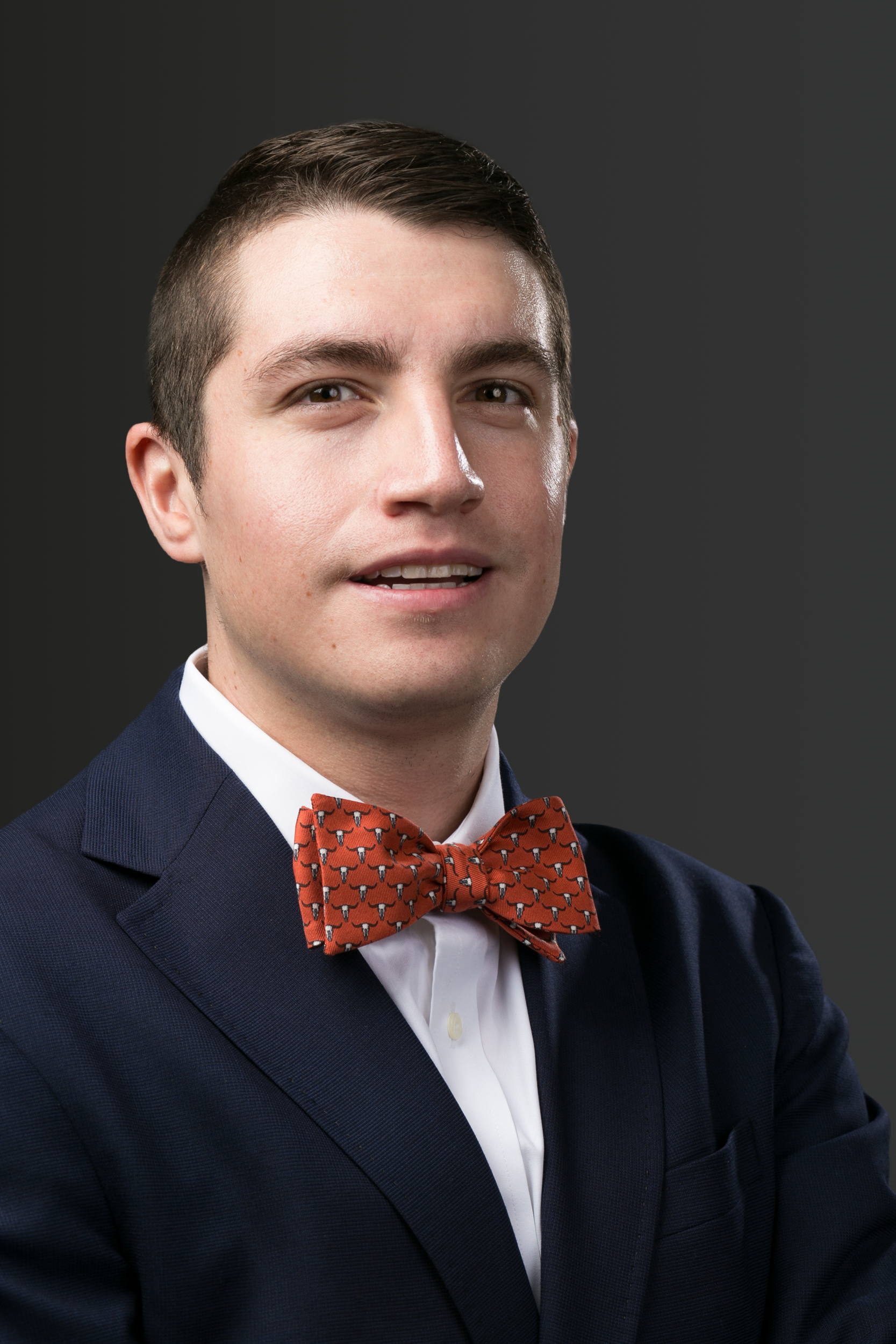 A person wearing a blue patterned blazer, white collared shirt, and orange polka-dot bowtie against a dark background.