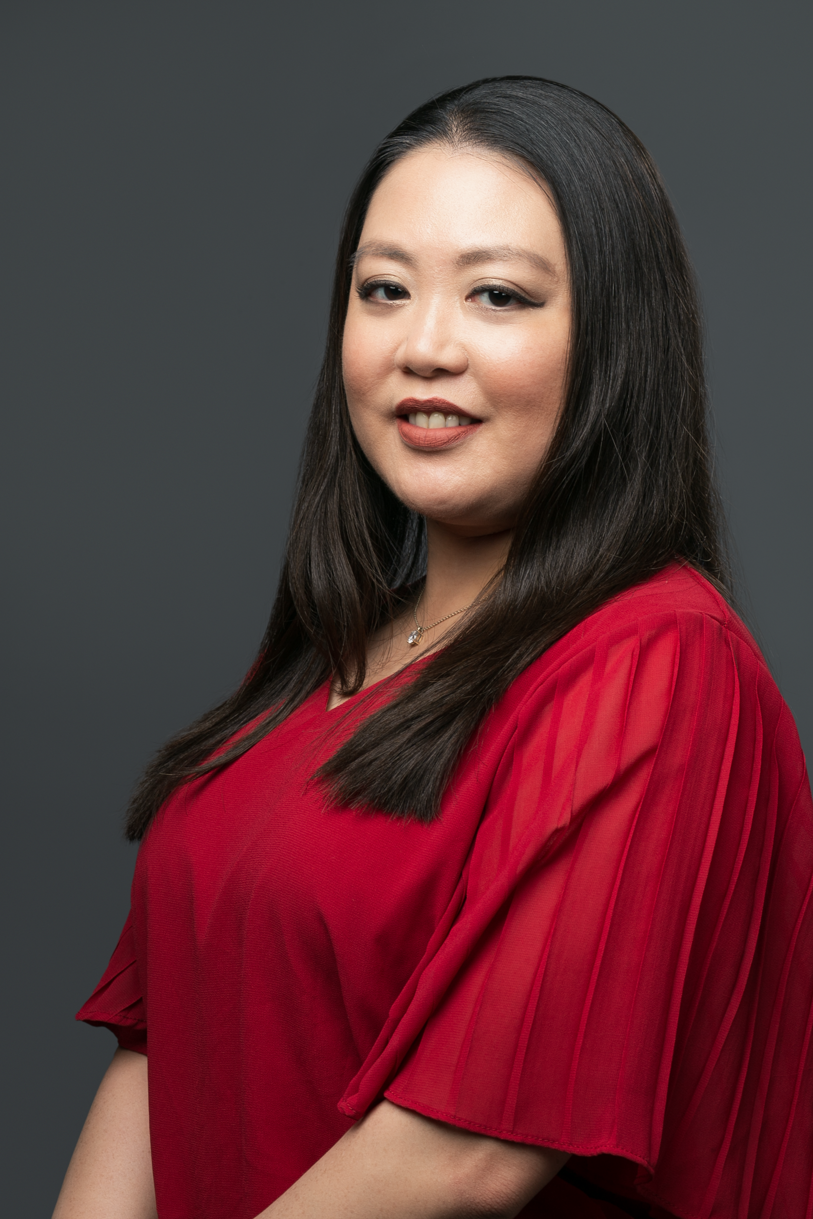 A person with long dark hair smiles gently against a solid grey background, wearing a red top with a textured finish.