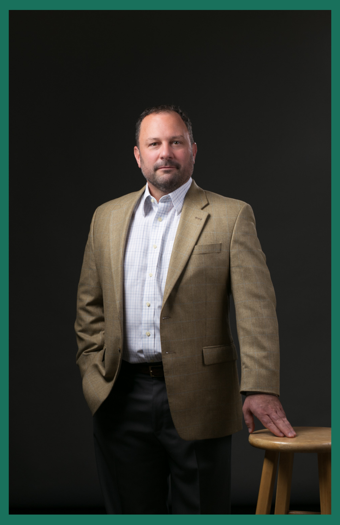 Man in blazer, hand in pocket, leaning on a stool, against a gray backdrop.