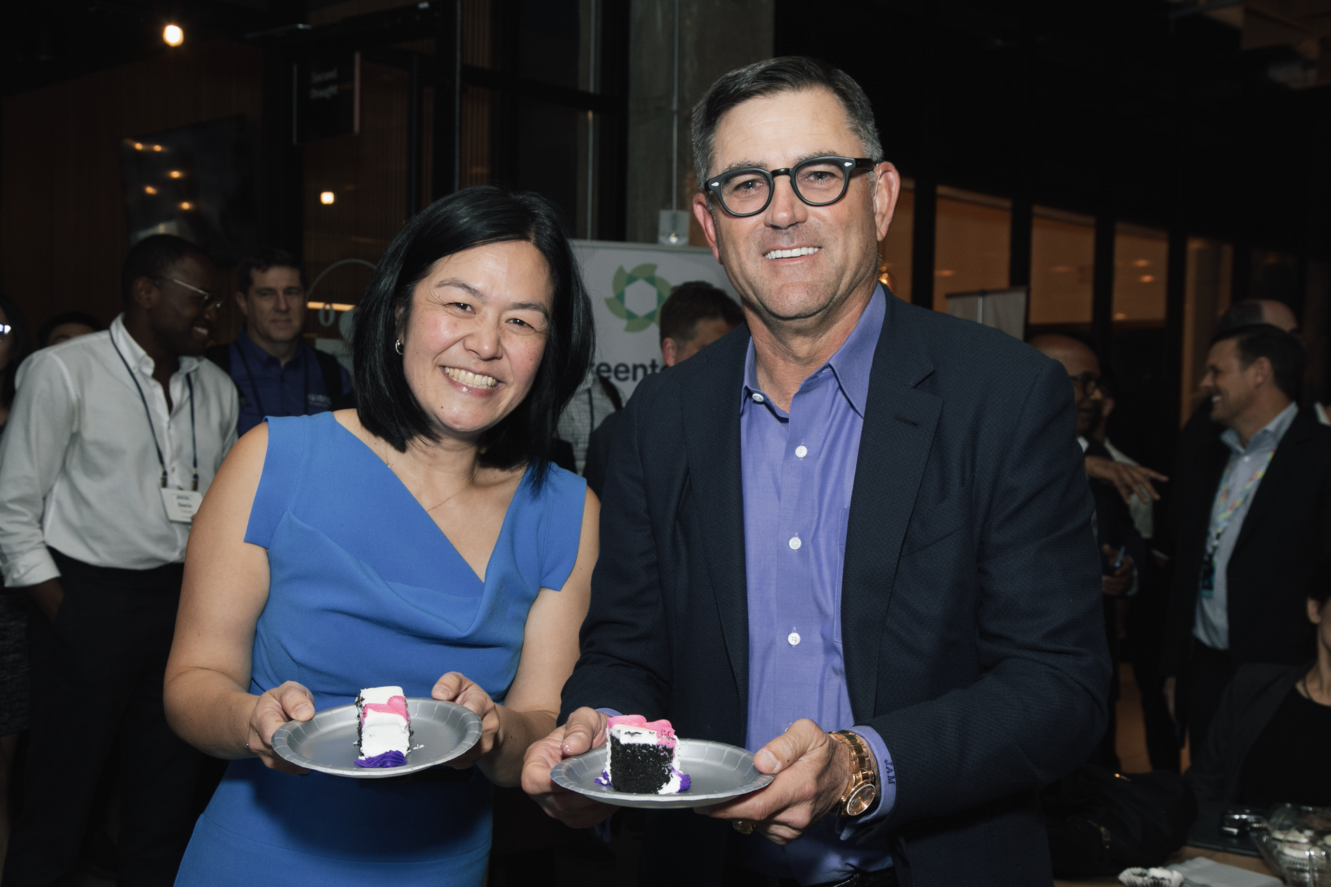 Two smiling people hold small plates with cake slices at an indoor event, with others visible in the blurred background.