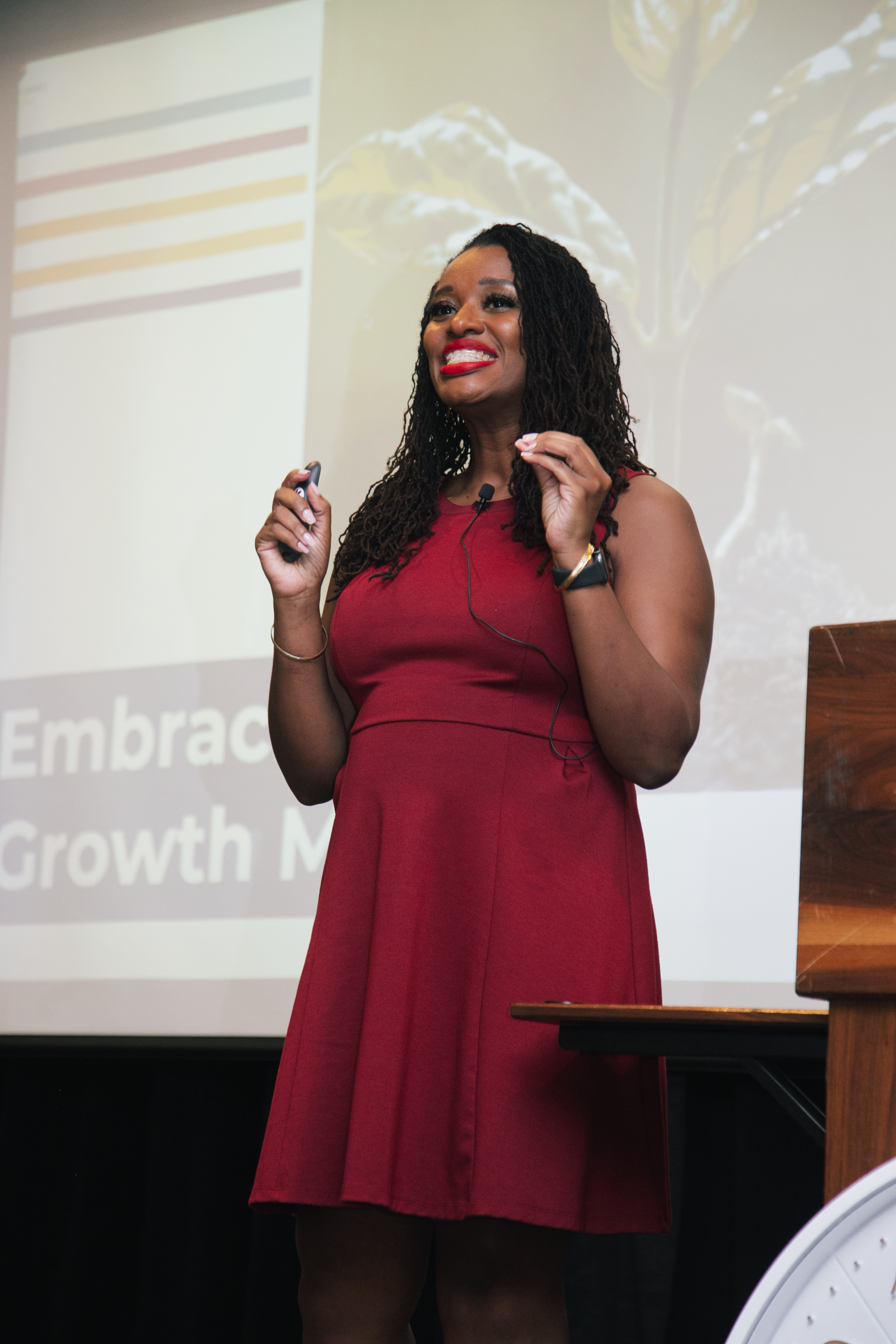 A person in a red dress speaks at a presentation, gesturing with their hands in front of a Growth Mindset slide.