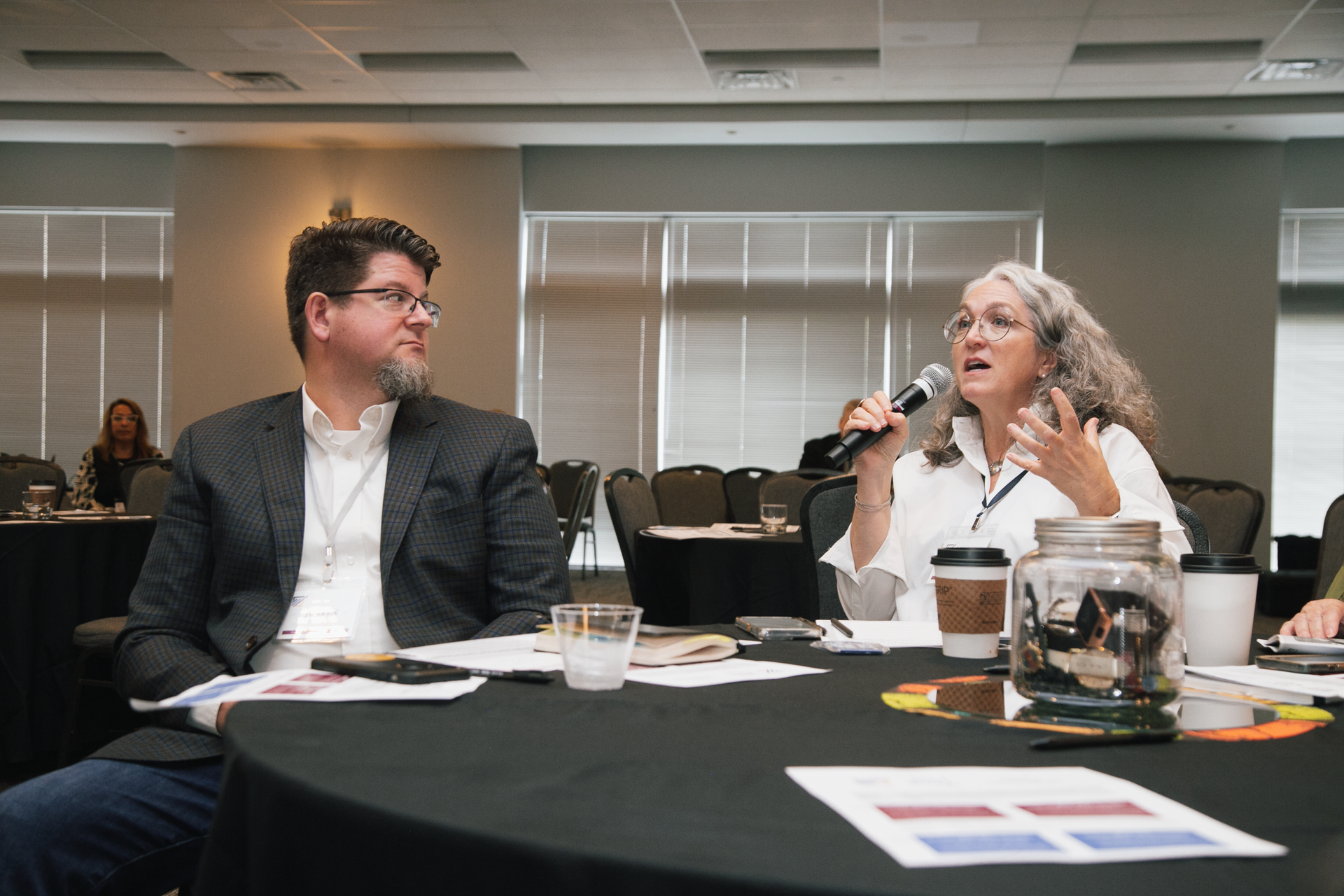 A person speaks into a microphone at a conference table, while a person in a blazer listens attentively.