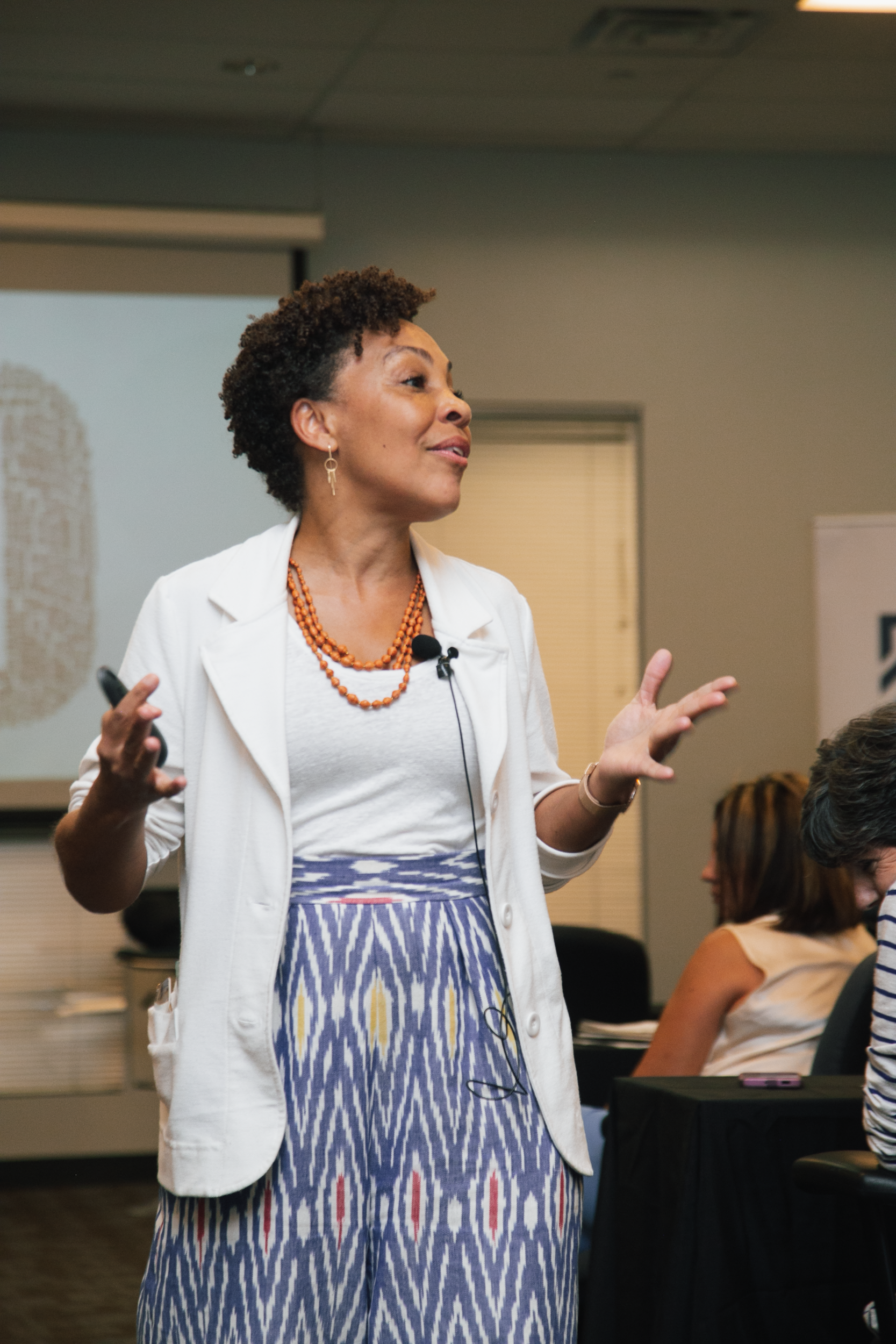 A speaker in a white blazer and patterned blue skirt gestures while presenting in a room with a projector screen.