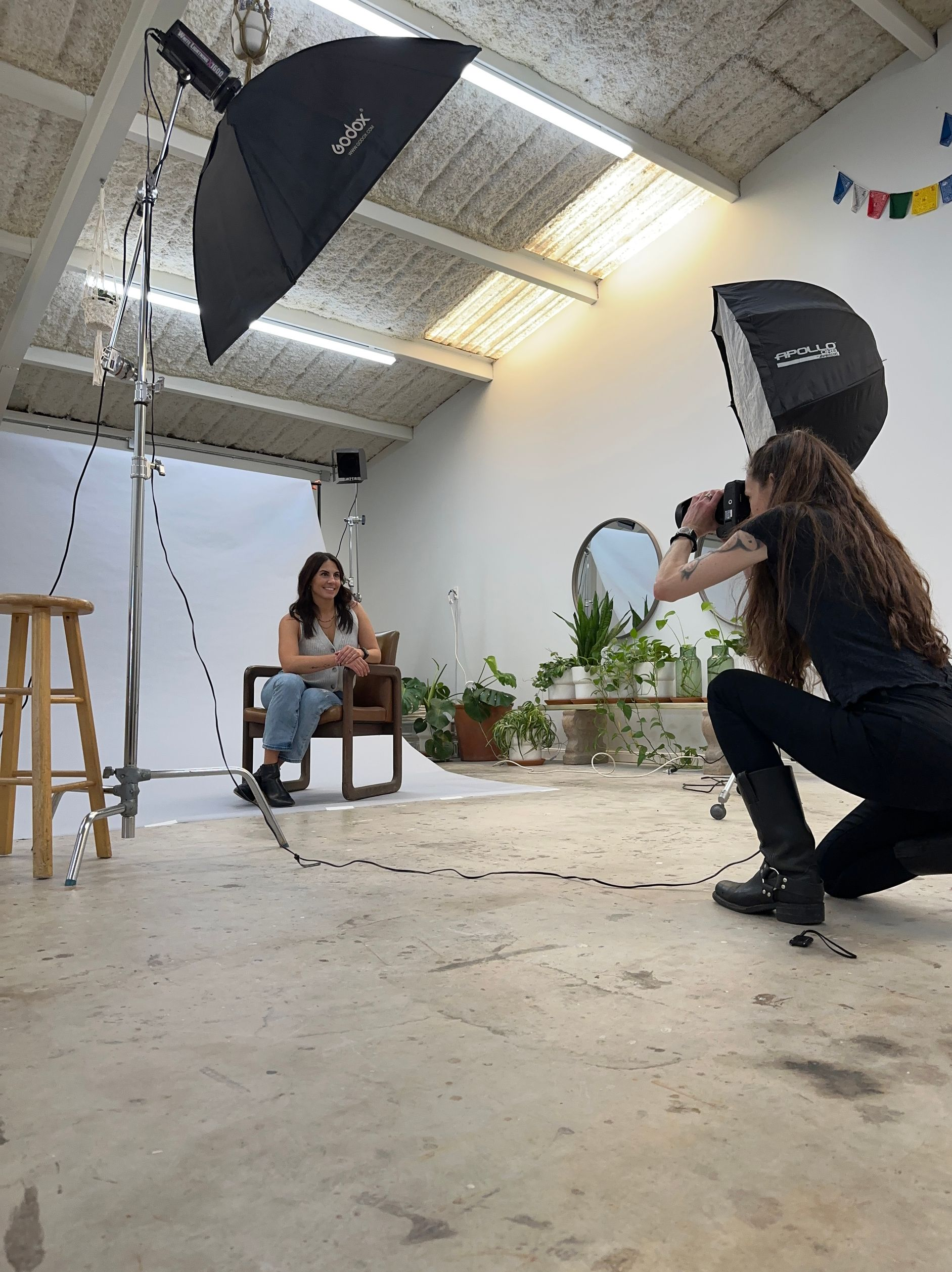 Photographer kneels, taking photos of a man sitting on a stool in a studio with lighting equipment.