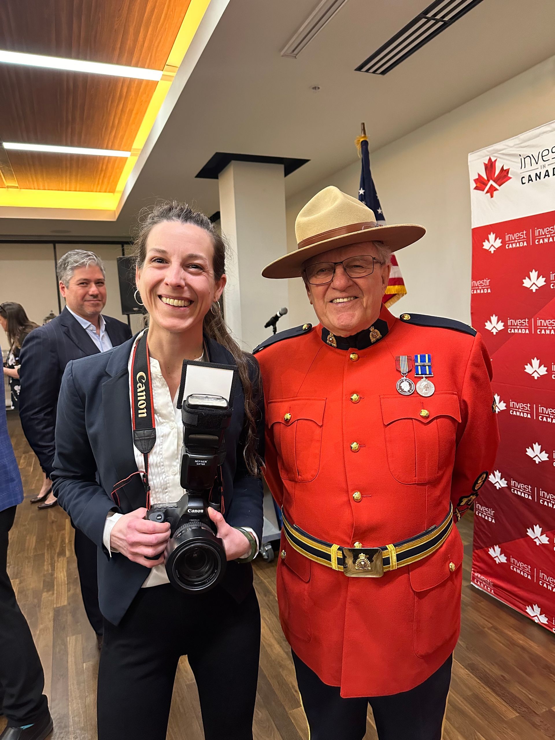Woman holding large camera posing with RCMP officer in red serge indoors at event backdrop