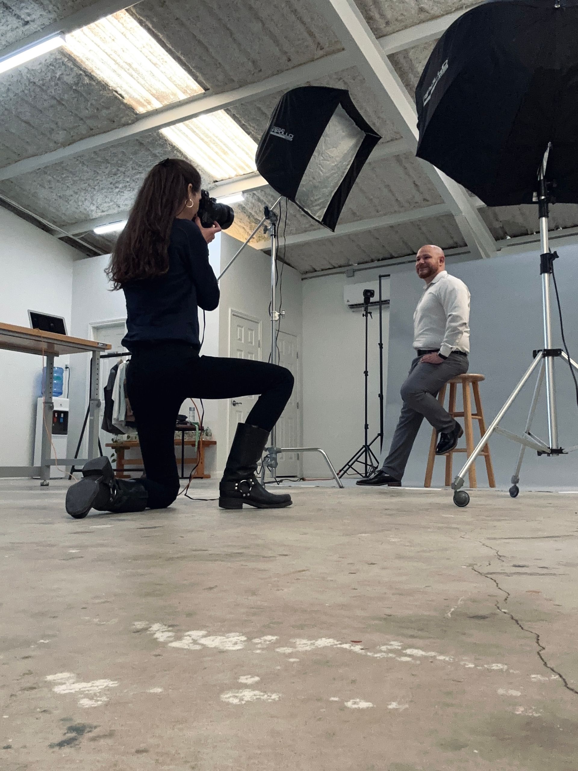 Photographer kneeling, taking photo of a person sitting on chair in studio. Two softboxes illuminate the subject.