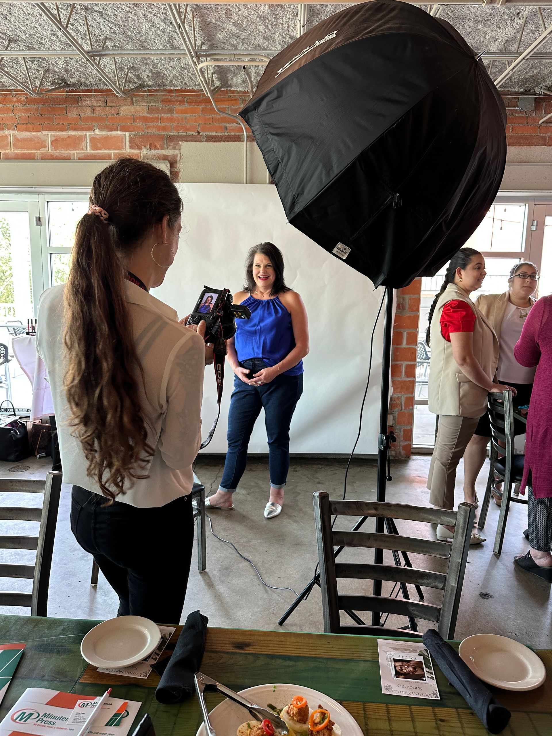 Indoor photo shoot setup with large softbox lights, people posing near a table in a brick-walled room