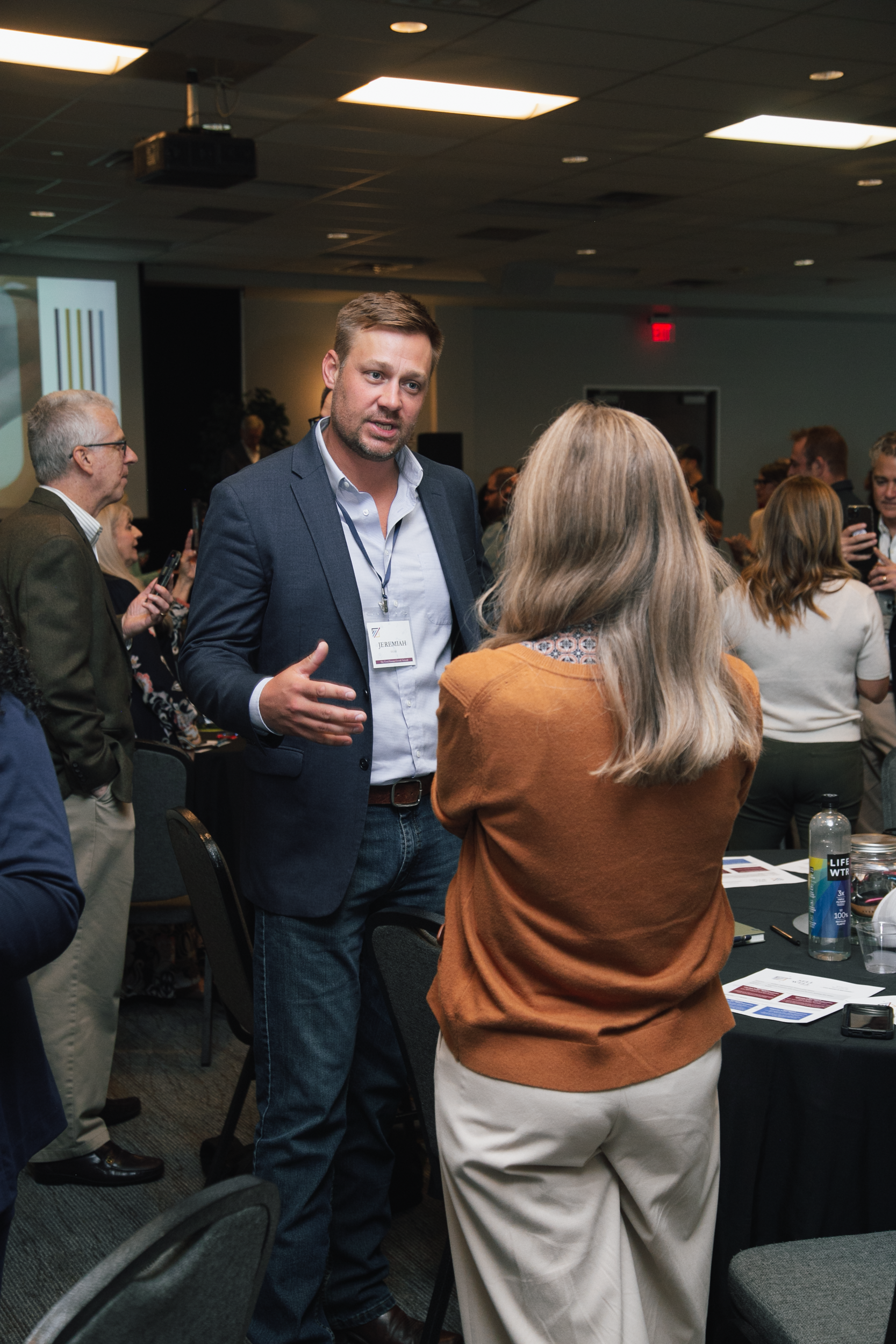 A man in a navy blazer speaks to someone wearing an orange sweater at a professional networking event.