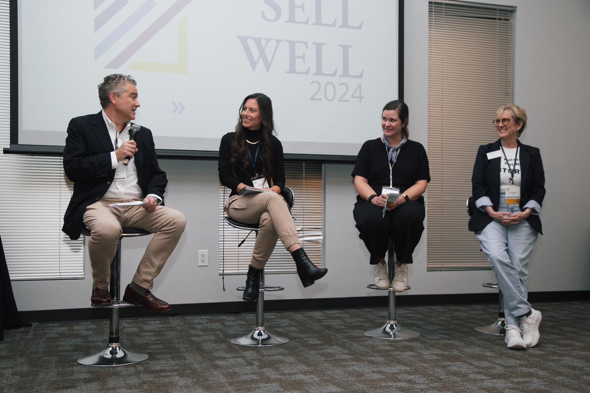 Four people sit on tall stools on a stage in front of a screen reading 