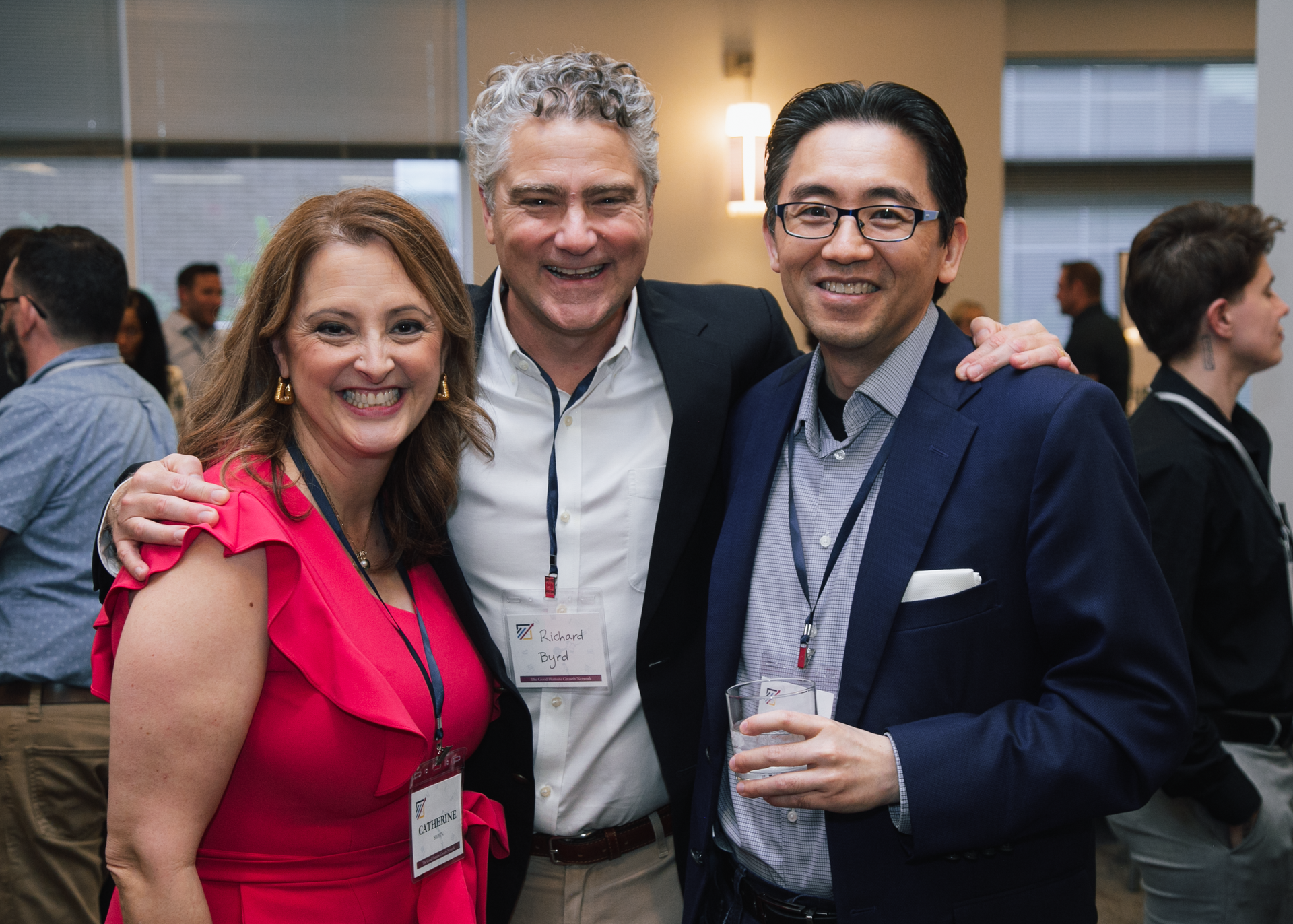 Three people smiling at a networking event. They wear conference badges, and the middle person has an arm around the others.