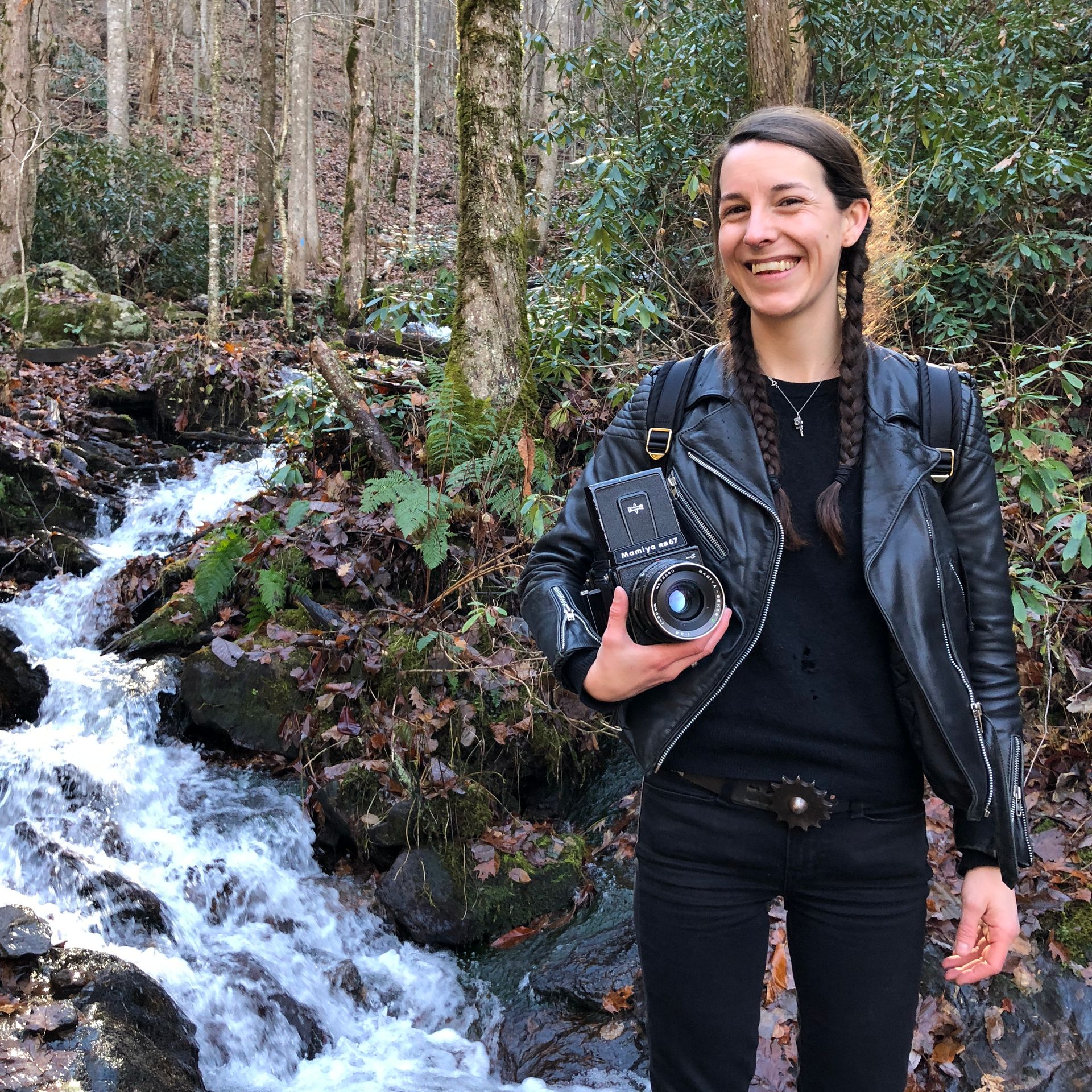 Smiling person holding a camera beside a forest waterfall