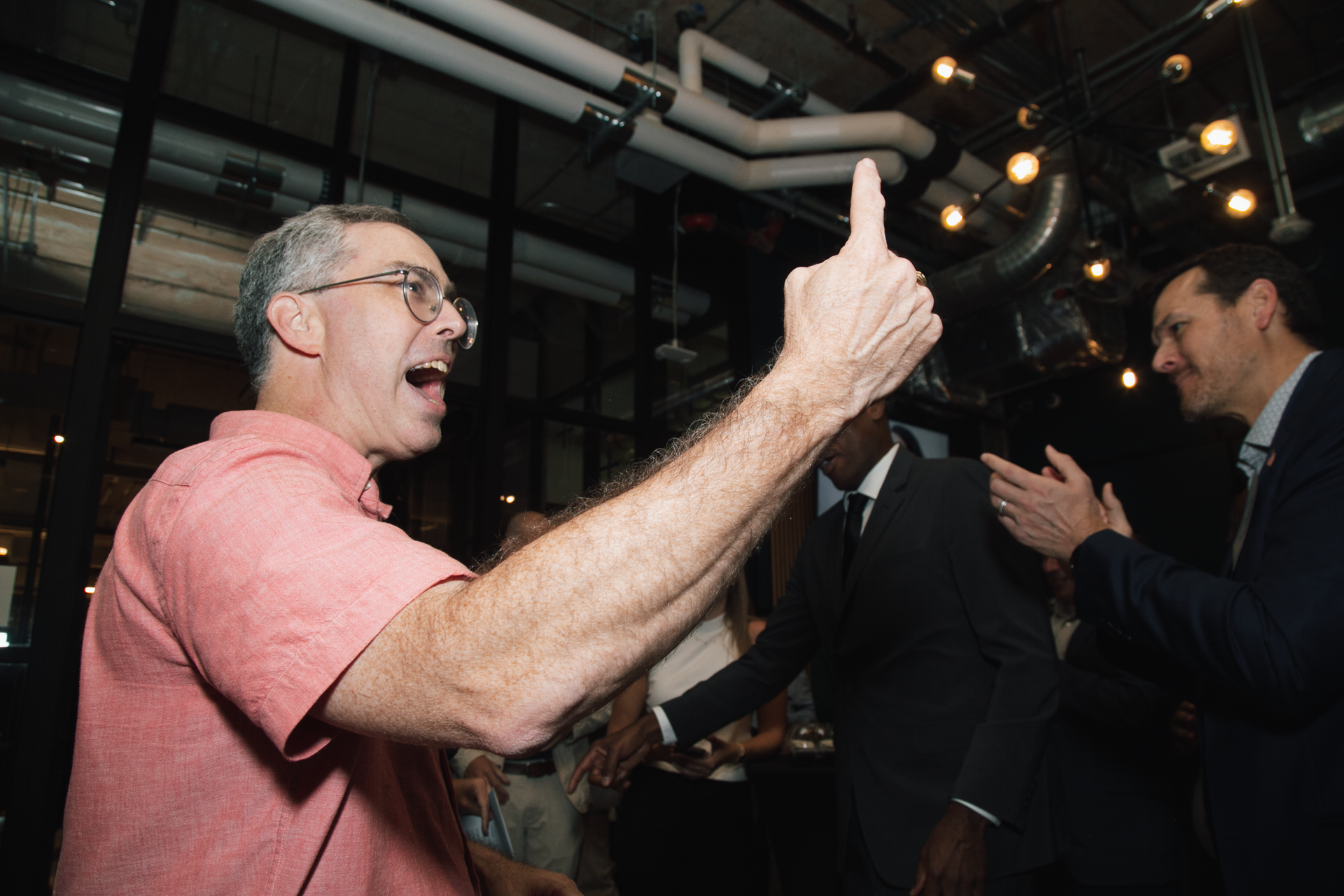 A person in a pink shirt excitedly points a finger upward in a dim, industrial-style room while others watch and clap.