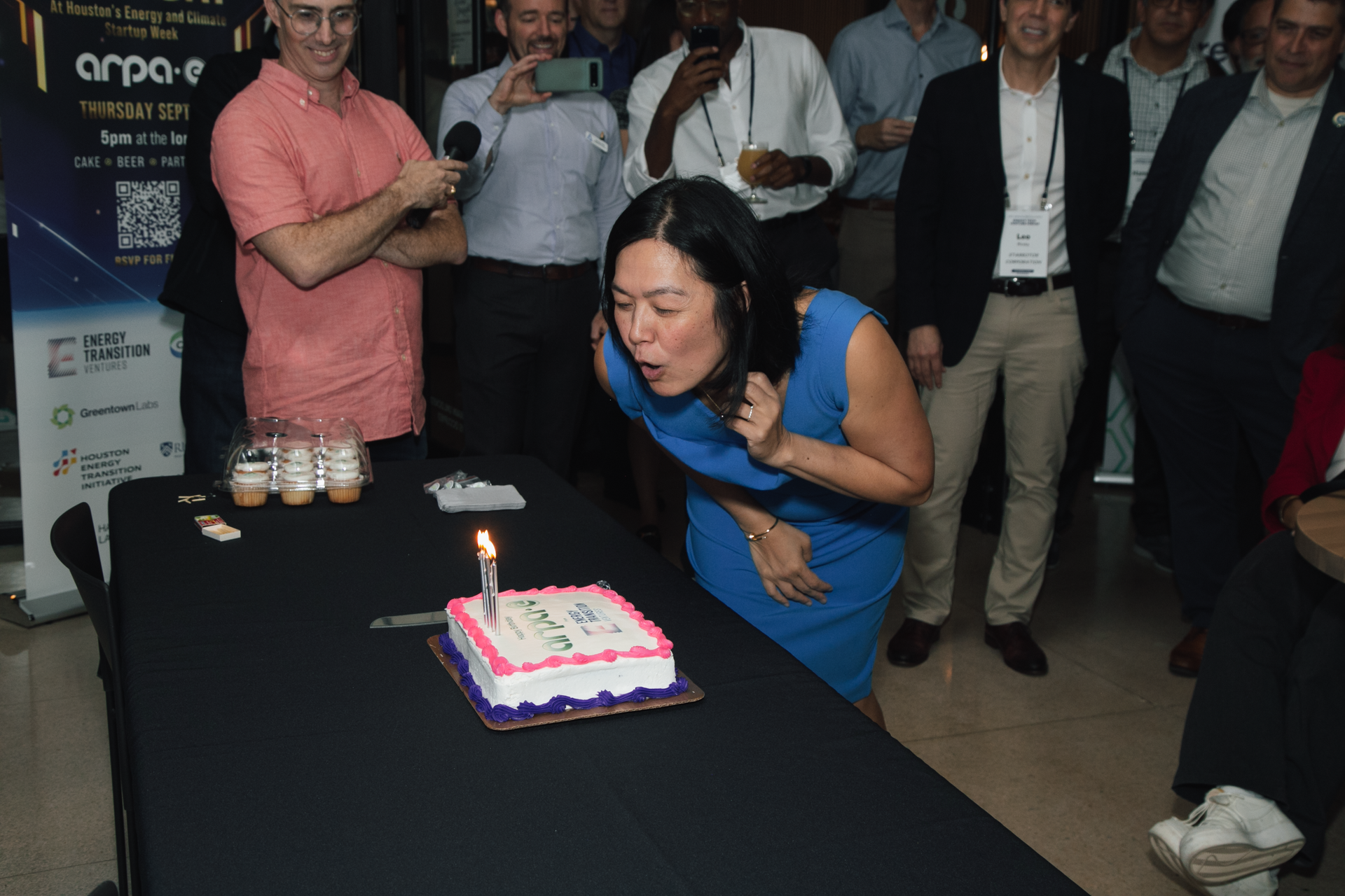 A person in a blue dress leans over a table to blow out a single lit candle on a cake, surrounded by an observant group.