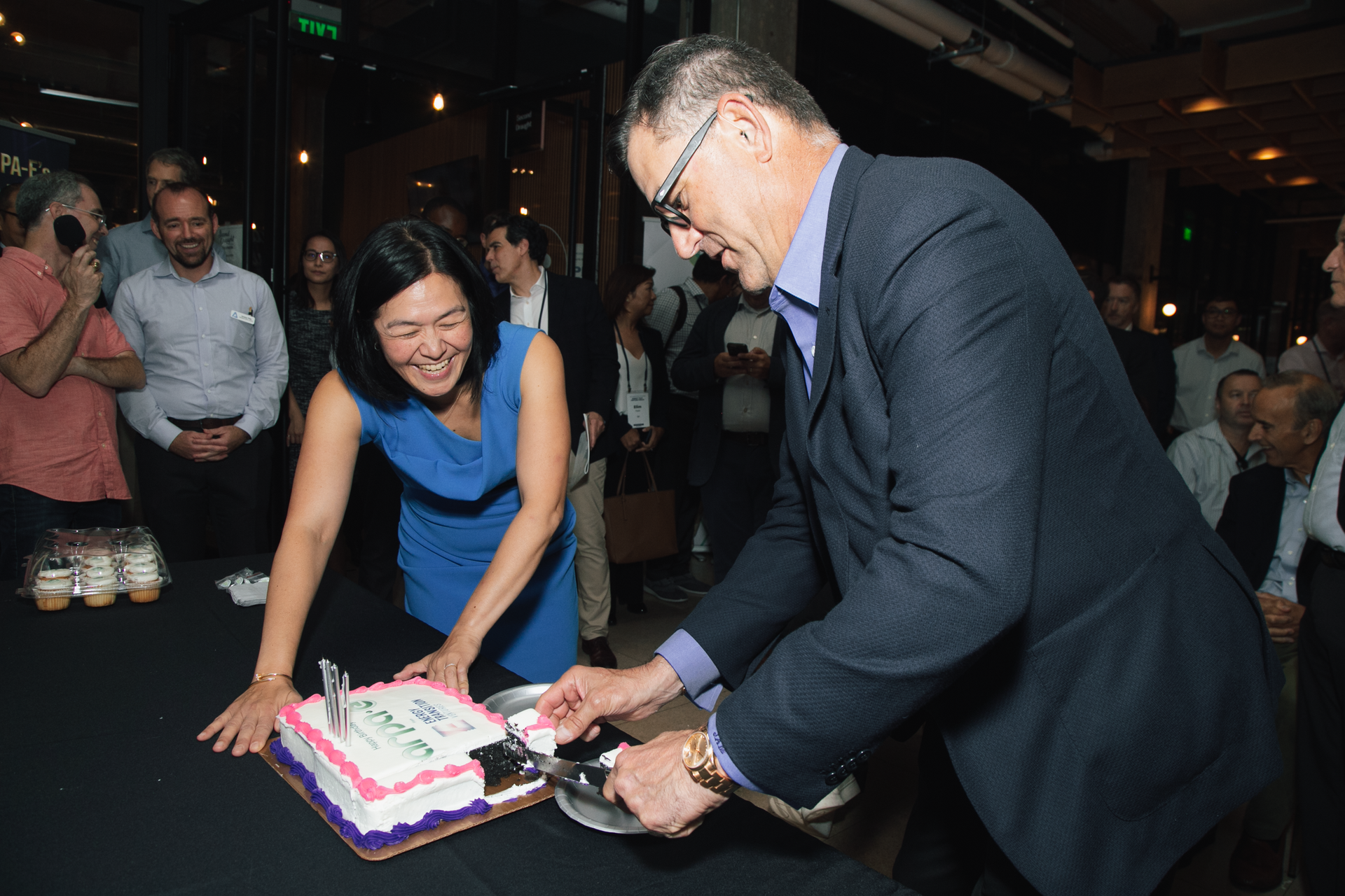 Two people cut a rectangular cake at an indoor event, both smiling as they serve the dessert to guests nearby.
