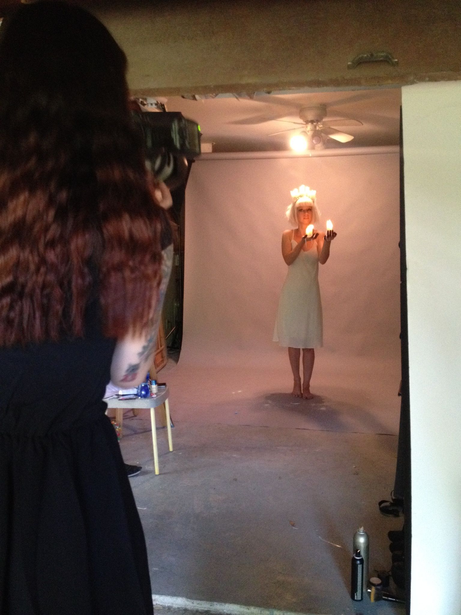 Person in a white dress stands on a small stage under a ceiling fan, holding a candle in a dim room.