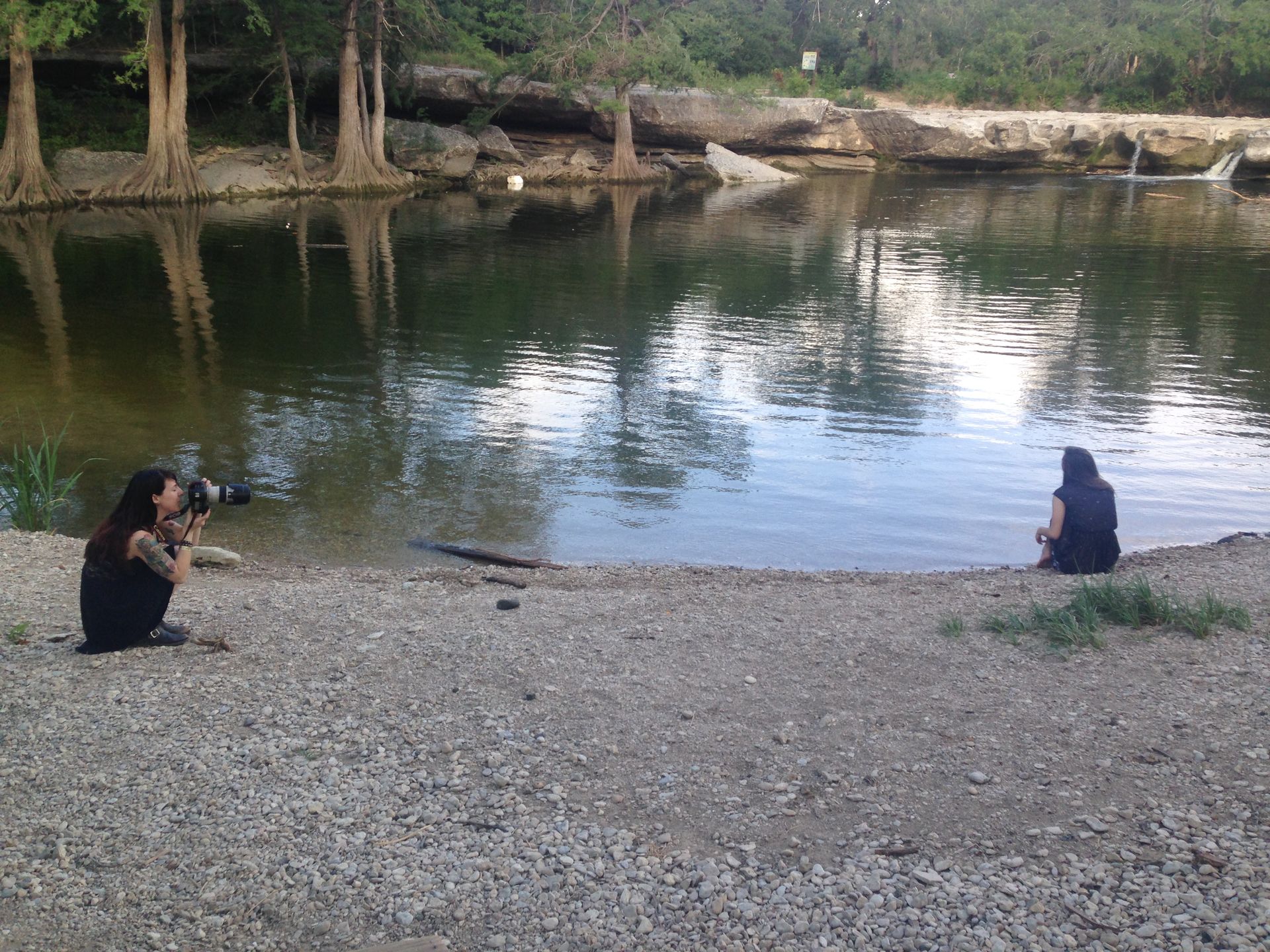 Two people sit by a calm river on a rocky shore, one taking a photo across the water.