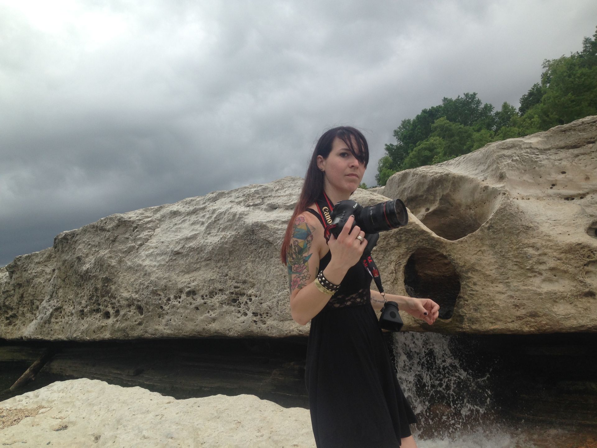 Person holding a camera beside a large rock with a small waterfall under cloudy skies