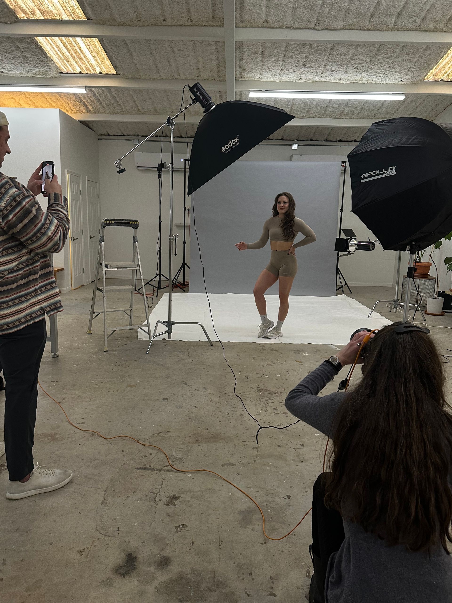 Studio photo shoot with model posing on a white backdrop, lit by softbox lights and photographed by crew
