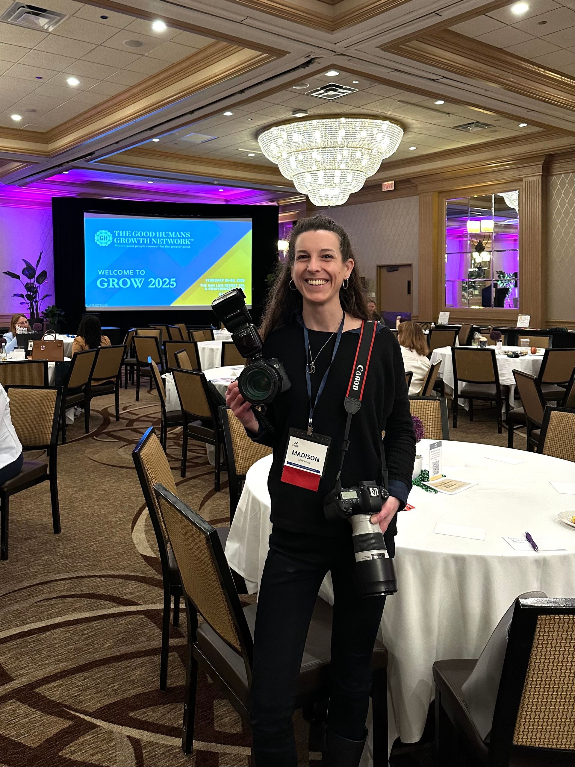 Woman standing in a banquet hall beside round tables, smiling with a camera and lanyard, near a presentation screen