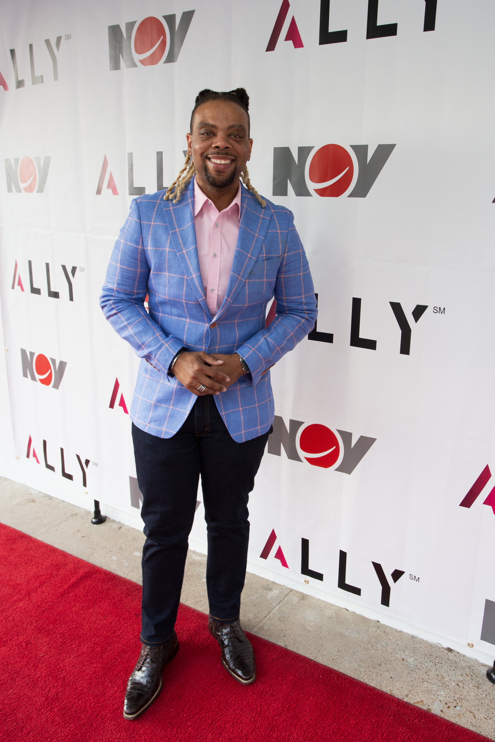A smiling person with long hair stands on a red carpet in front of a white step-and-repeat backdrop with red logos.