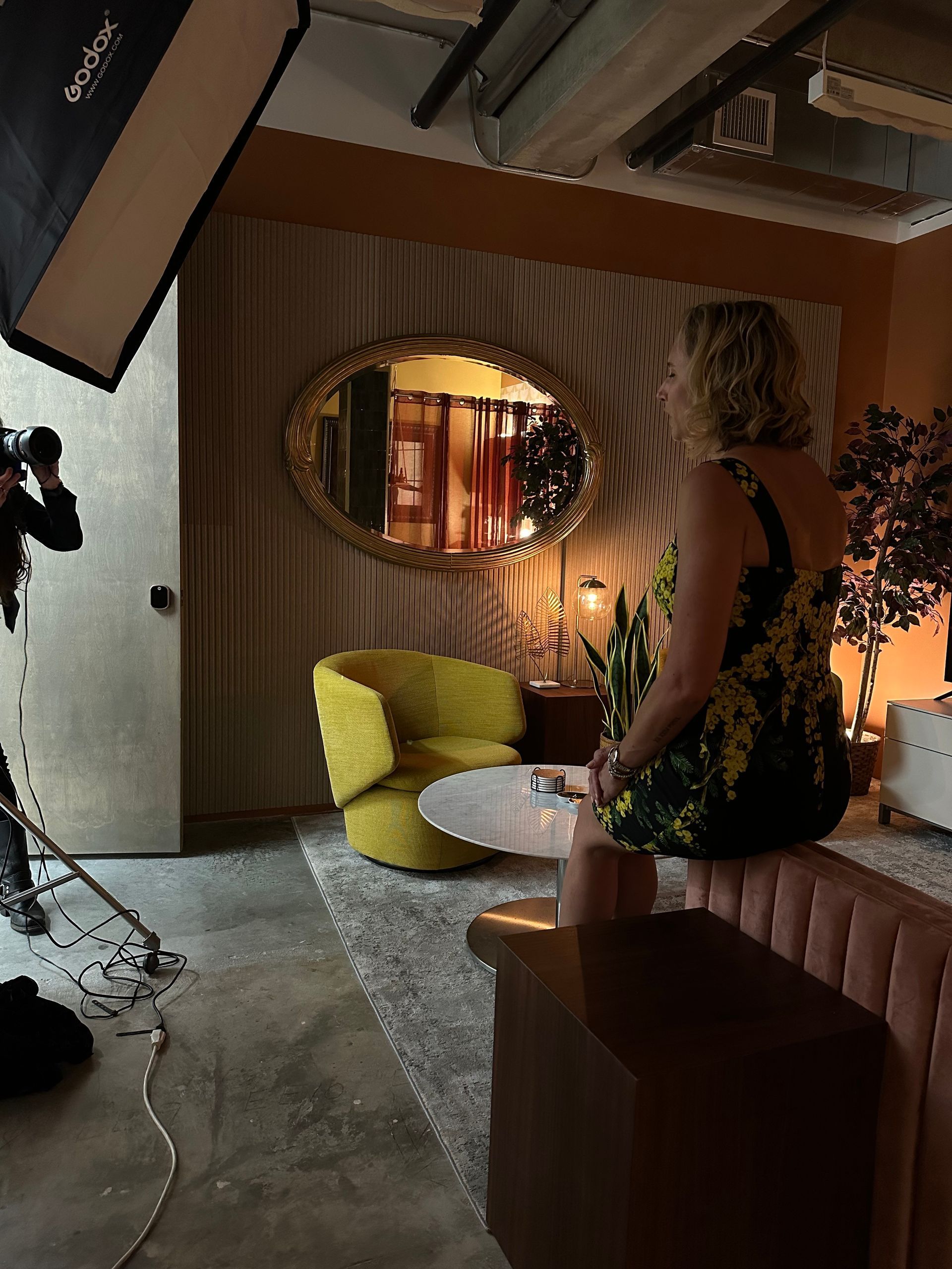 Cozy lounge with yellow chair, round mirror, striped wall, warm lighting, and a standing person near a couch