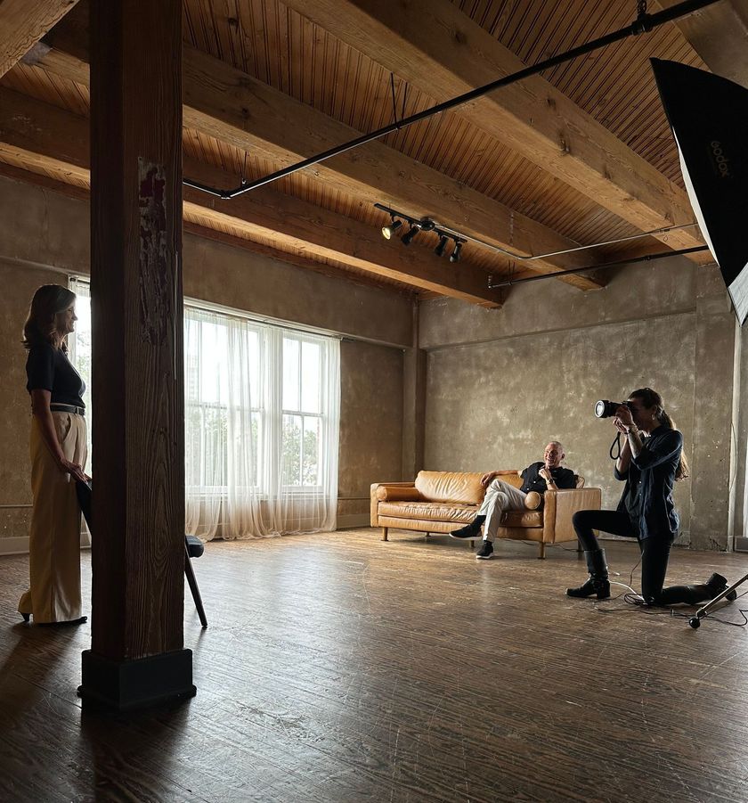 A photographer kneels in a studio to take a picture of a person, while another person sits on a leather sofa nearby.