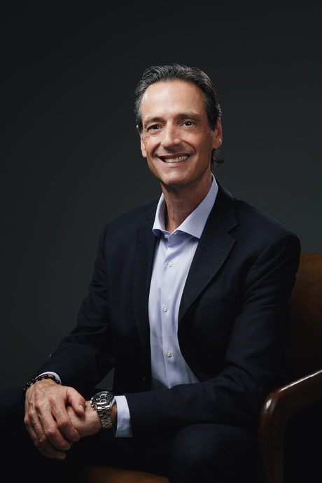 Man in dark blazer and white shirt seated against a dark background, smiling at the camera.