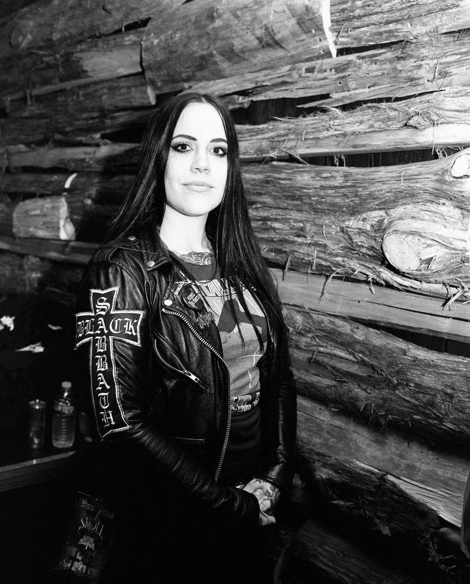 Person with long dark hair and leather jacket seated against a rustic wooden log wall.Shot on medium format film, and then developed and printed in a traditional wet darkroom by the photographer,  Madison Avery Somerville.