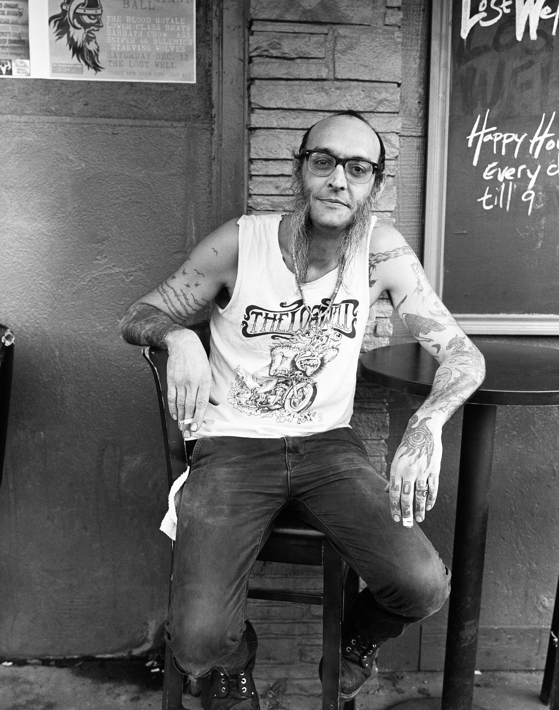Man sitting outside a doorway, wearing glasses and a graphic T-shirt, with tattooed arms. Shot on medium format black and white film by Madison Avery Somerville. Developed and printed in a traditional wet darkroom by the photographer.