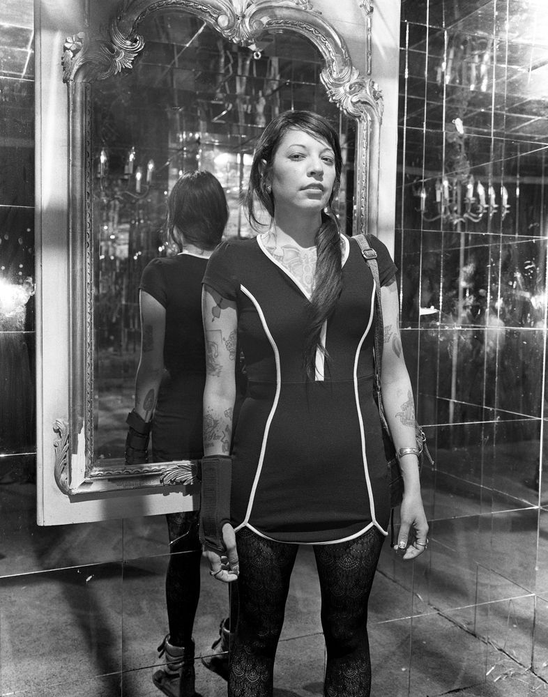Woman standing in a mirrored hallway, wearing a dark dress and leggings, with reflections behind her. Shot on medium format film by Madison Avery Somerville. Developed and printed in a traditional wet darkroom by the photographer.