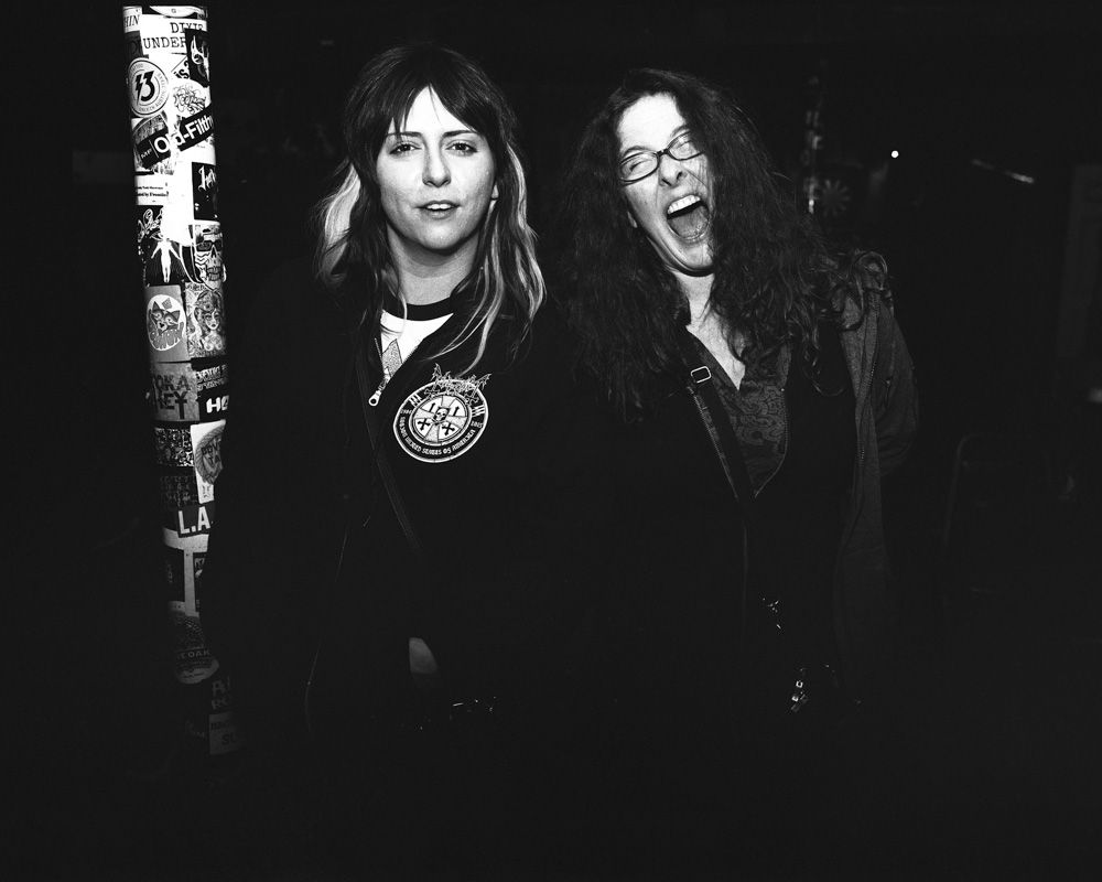 Two people in dark clothing laughing in a dark bar beside a poster-covered pole. Shot on medium format film. Developed and printed in a traditional wet darkroom by the photographer, Madison Avery Somerville.