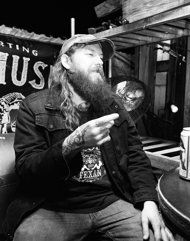 Bearded person in a cap and dark jacket, sitting indoors and smoking near a bar sign. Shot on medium format film by Madison Avery Somerville. Developed and printed in a traditional wet darkroom by the photographer.