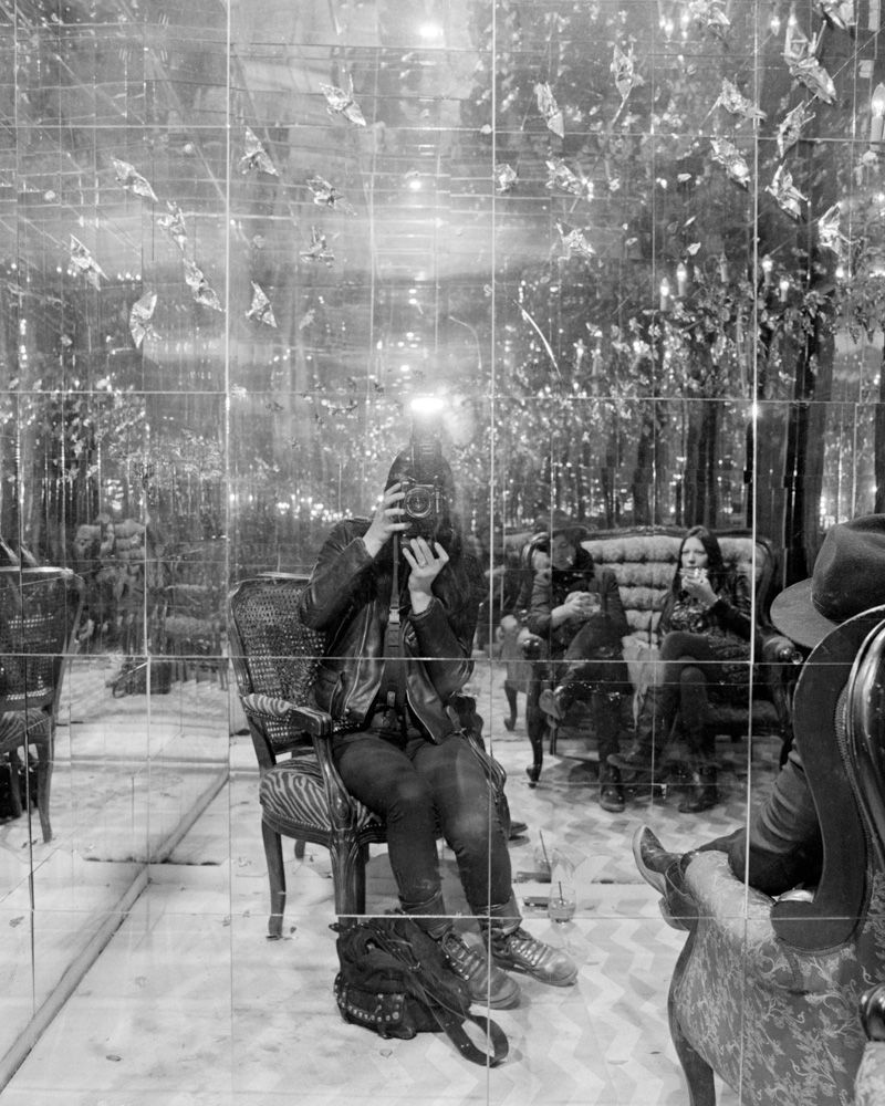 Person sitting on a chair in a glass-walled café, reflected by lights and trees outside. Shot on medium format film by Madison Avery Somerville. Developed and printed in a traditional wet darkroom by the photographer.