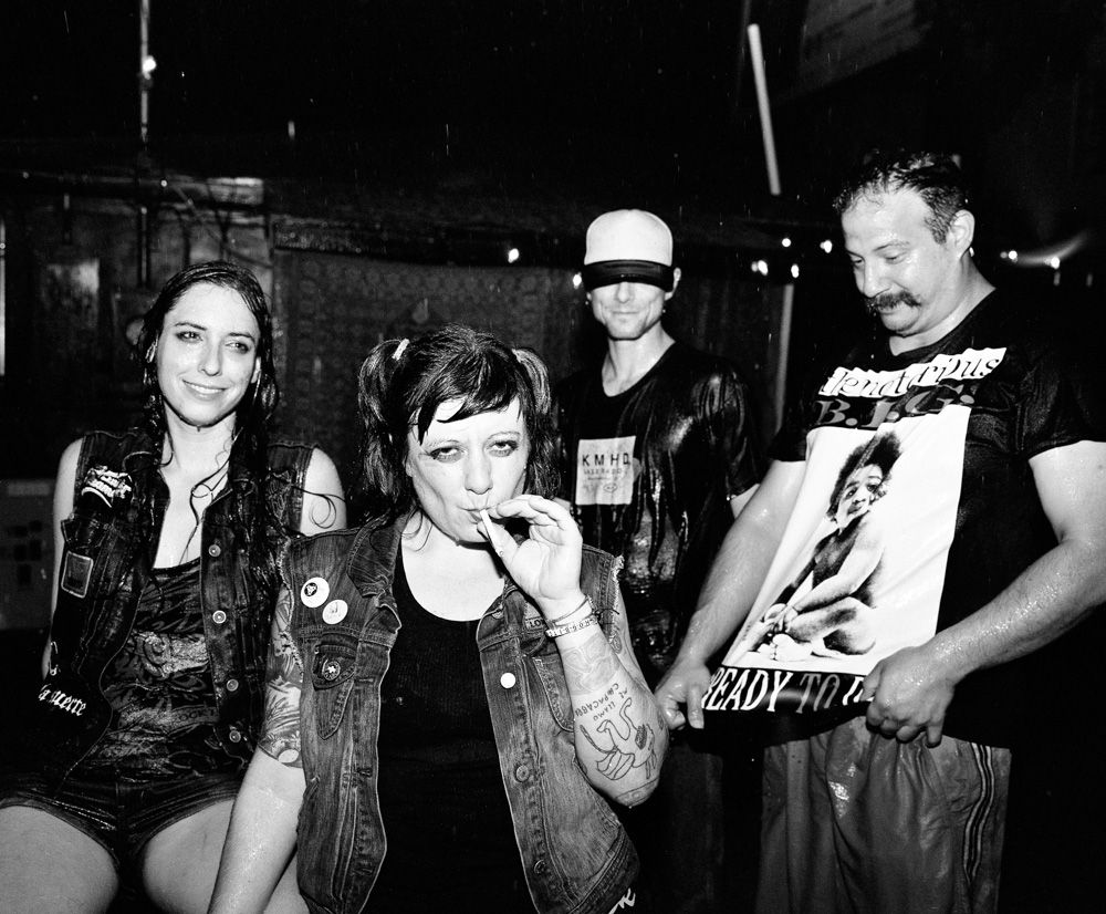 Black-and-white photo of four people at a concert, one smoking, in a dim venue. Shot on medium format film by Madison Avery Somerville. Developed and printed in a traditional wet darkroom by the photographer.